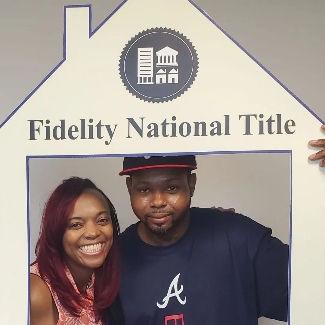 A man and a woman smiling behind a large cutout sign that reads 'Fidelity National Title' with a house-shaped top. The man is wearing a navy T-shirt with a white letter 'A' and a red and black cap. The woman has long, red hair and is wearing a patter