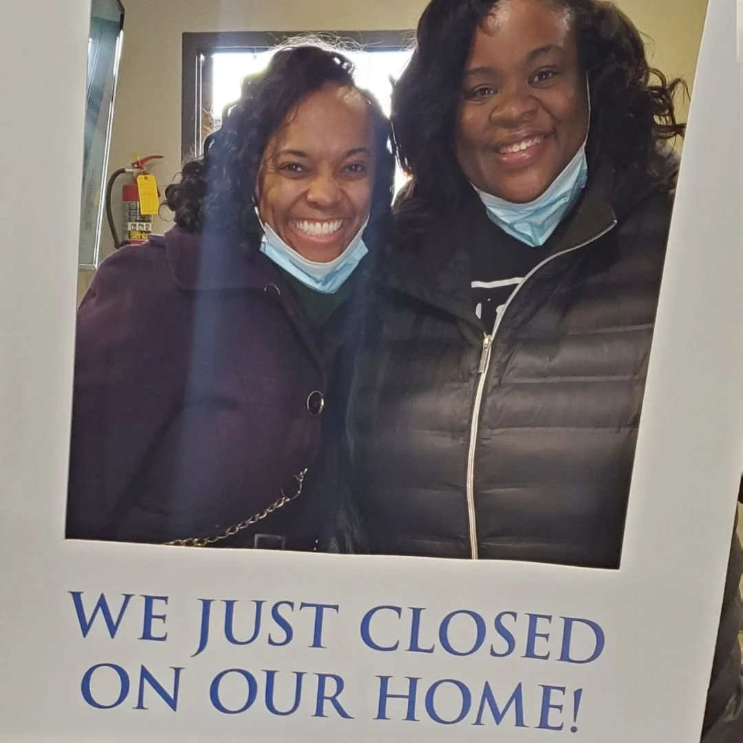 Two smiling women wearing face masks, holding a sign that reads 'We just closed on our home!'