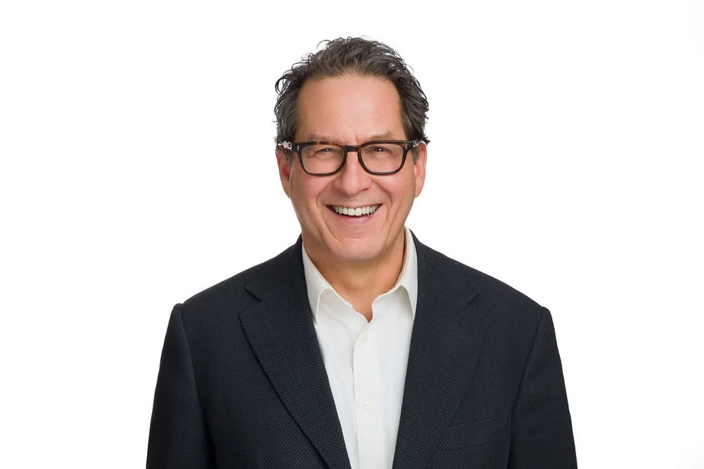 A middle-aged man with dark curly hair, glasses, and a bright smile, wearing a black blazer and white shirt, standing against a plain white background.