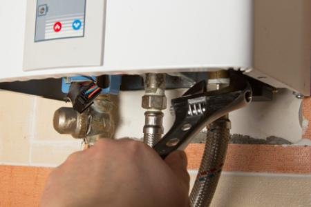 A person using a wrench to work on the plumbing connected to a white water heater or boiler mounted on a wall.