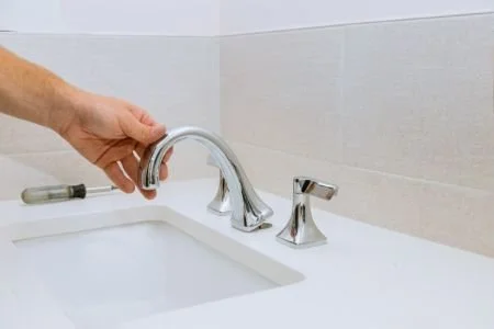 Person installing or adjusting a silver bathroom faucet in a white sink with beige tiled wall background.