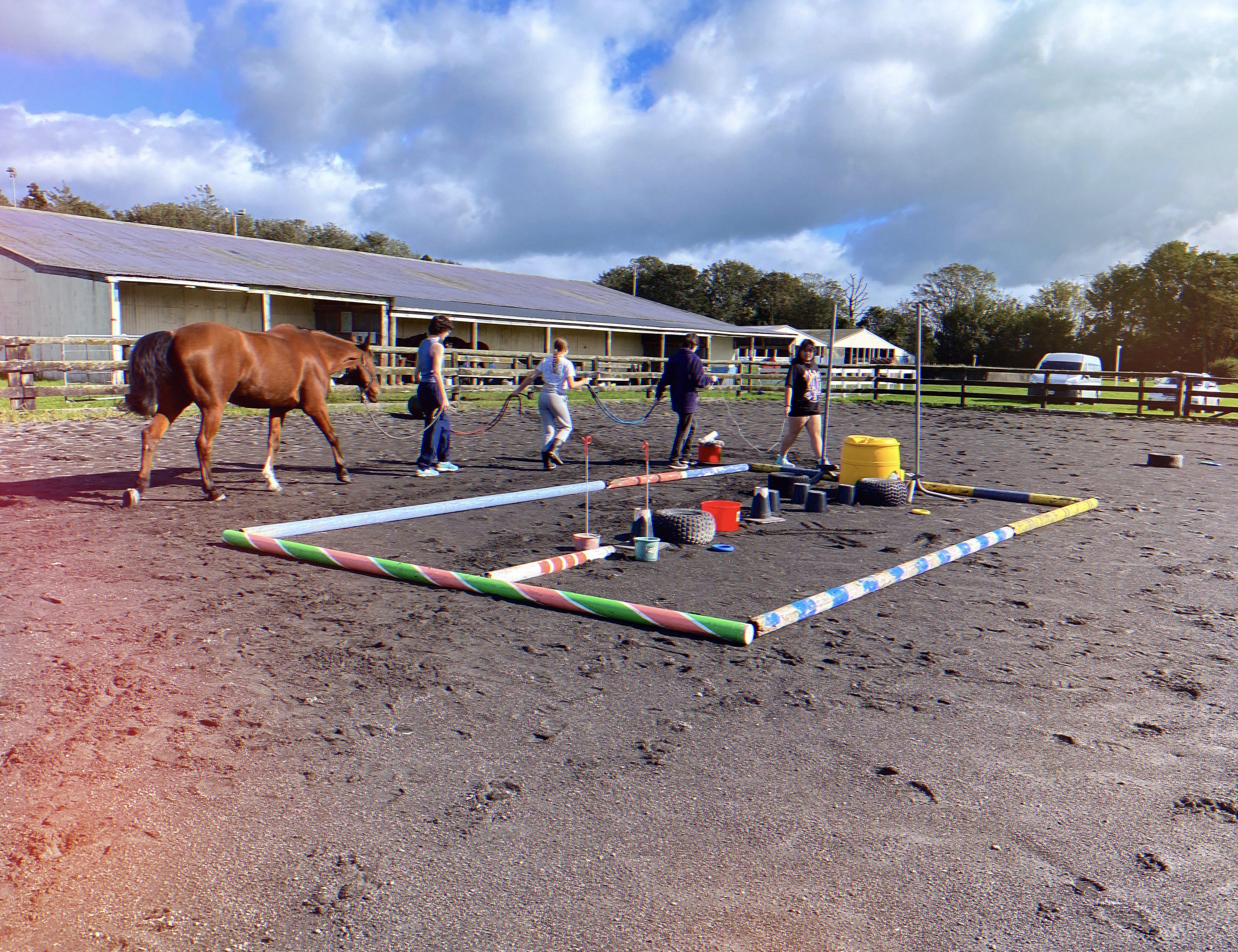 Multiple people walking a horse in a fenced outdoor arena on a sunny day with partly cloudy sky.