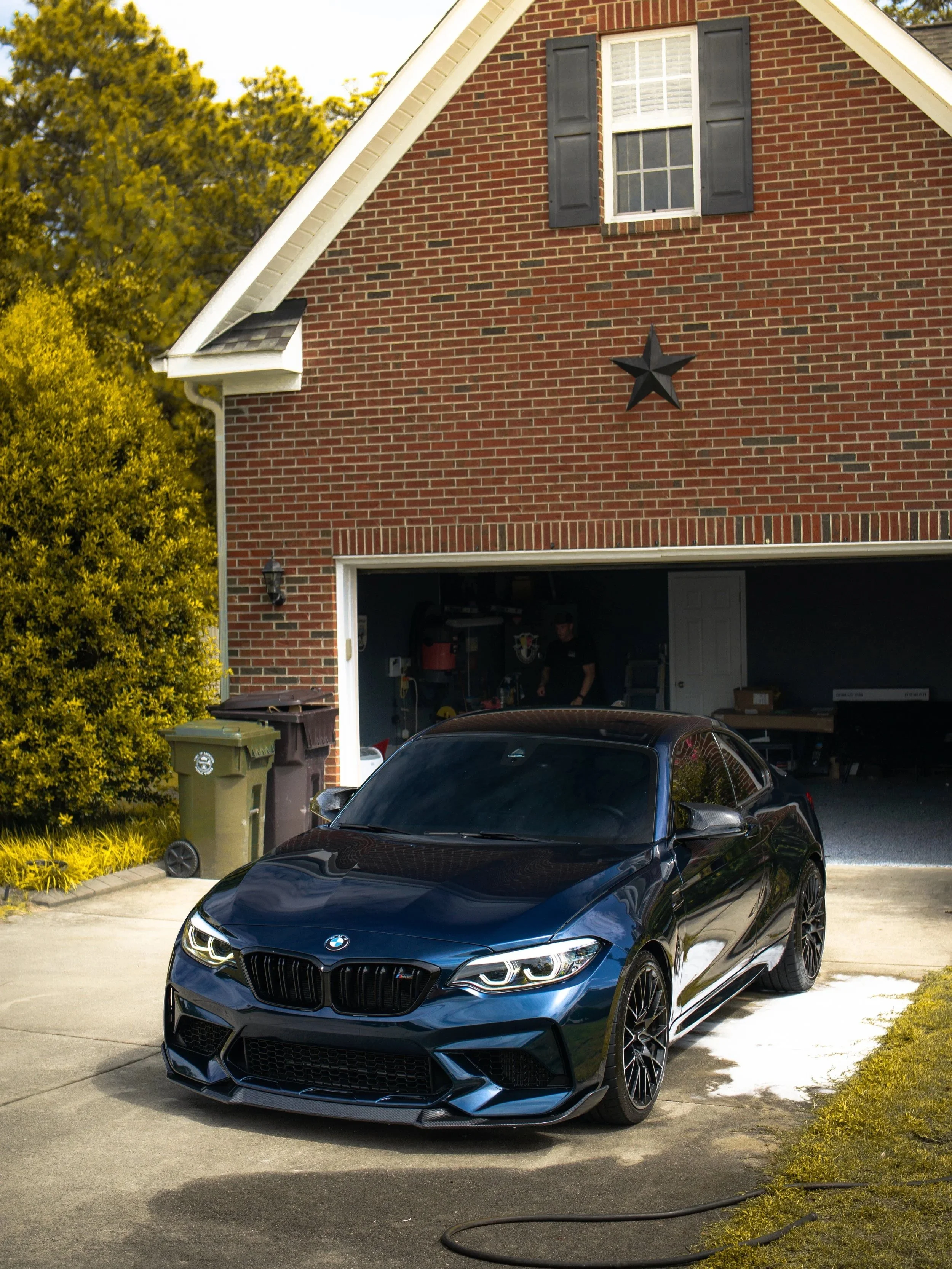 A black BMW M8 sports car parked in front of a red brick garage with an open door, showing a person inside working with tools. The garage has a star decoration on its exterior wall, a black shuttered window above, and surrounded by greenery and trash bins.