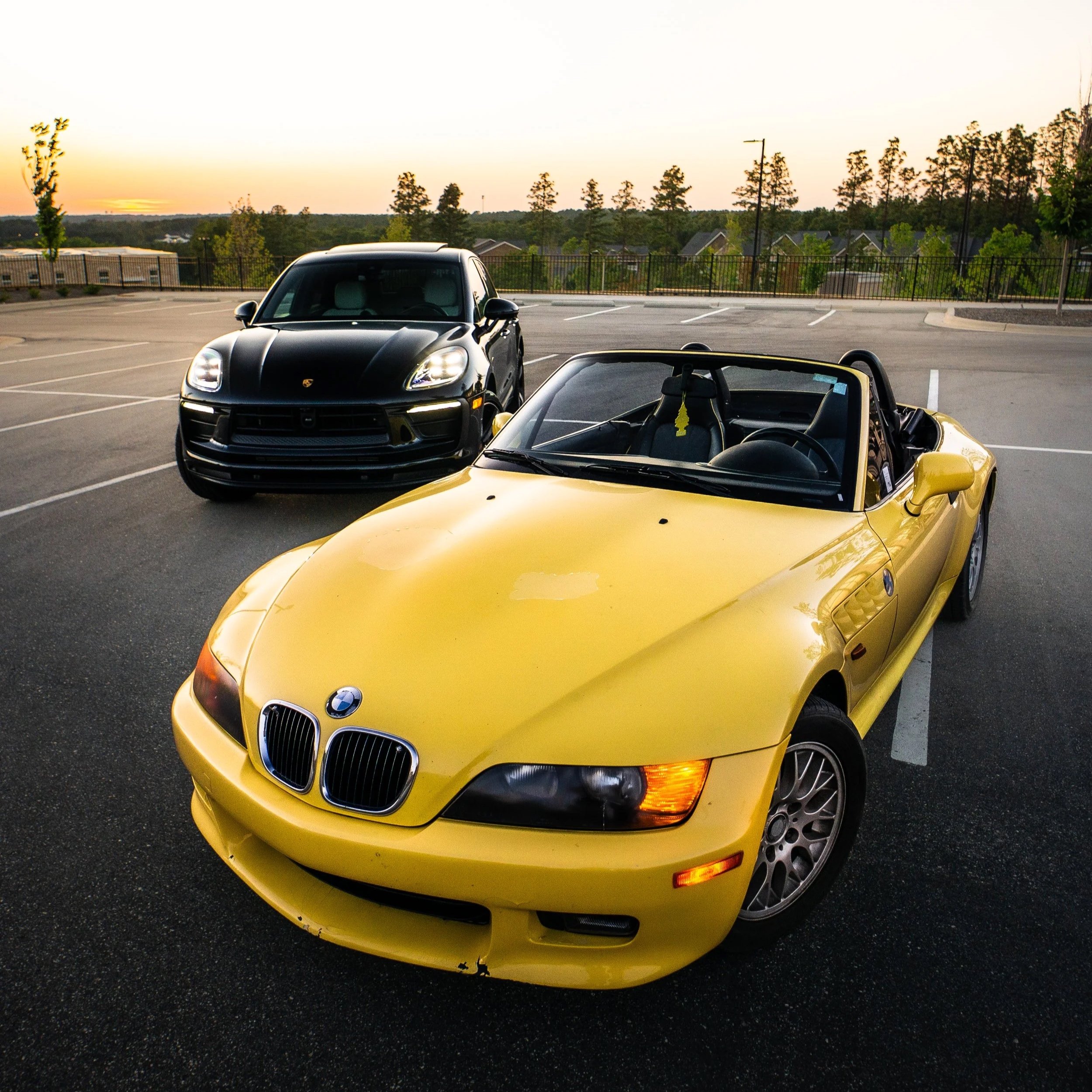 A yellow convertible sports car parked in an empty parking lot during sunset with a black Porsche SUV nearby and trees in the background.