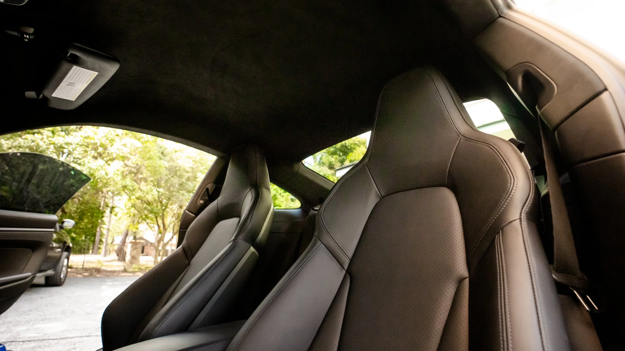 The interior of a car showing two black leather seats with a view outside through the windows, including trees and parked cars.
