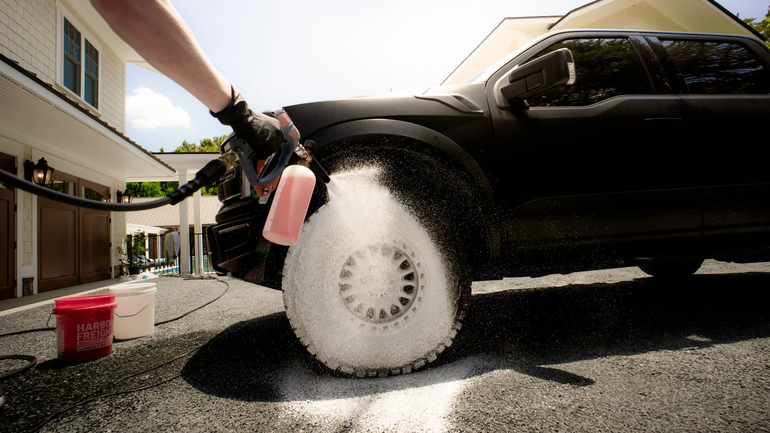 Person using a pressure washer to clean the tire of a black SUV on a driveway in front of a white house.