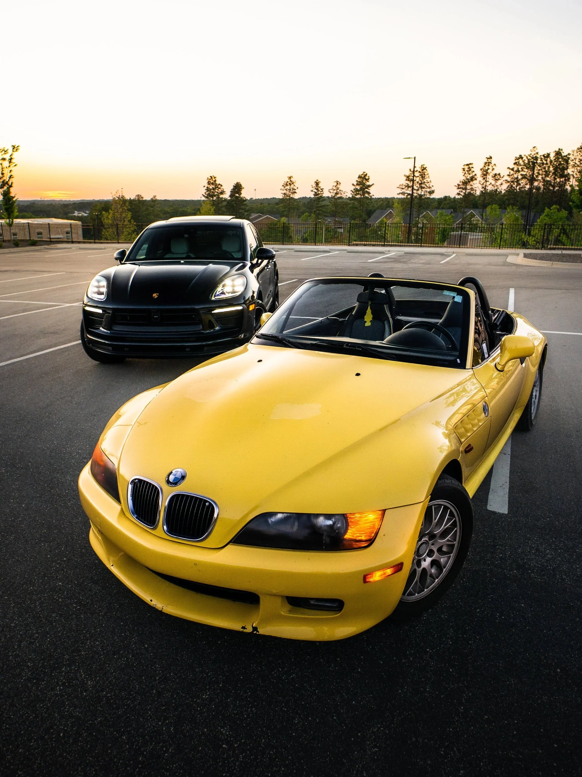 Yellow BMW convertible and black Porsche parked in an empty parking lot at sunset.