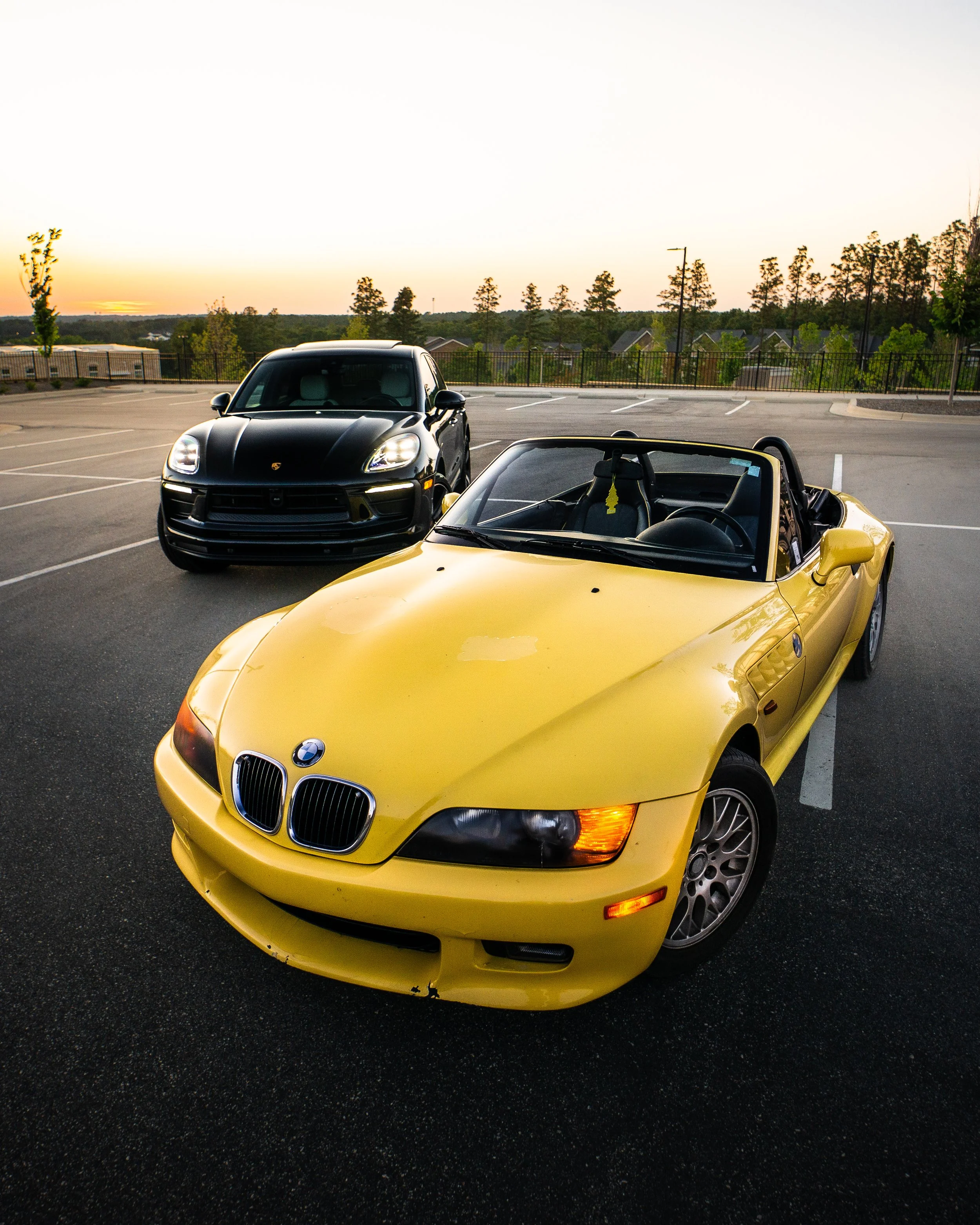 A yellow BMW Z3 convertible parked in front of a black Porsche Macan in an empty parking lot during sunset, with trees and houses in the background.