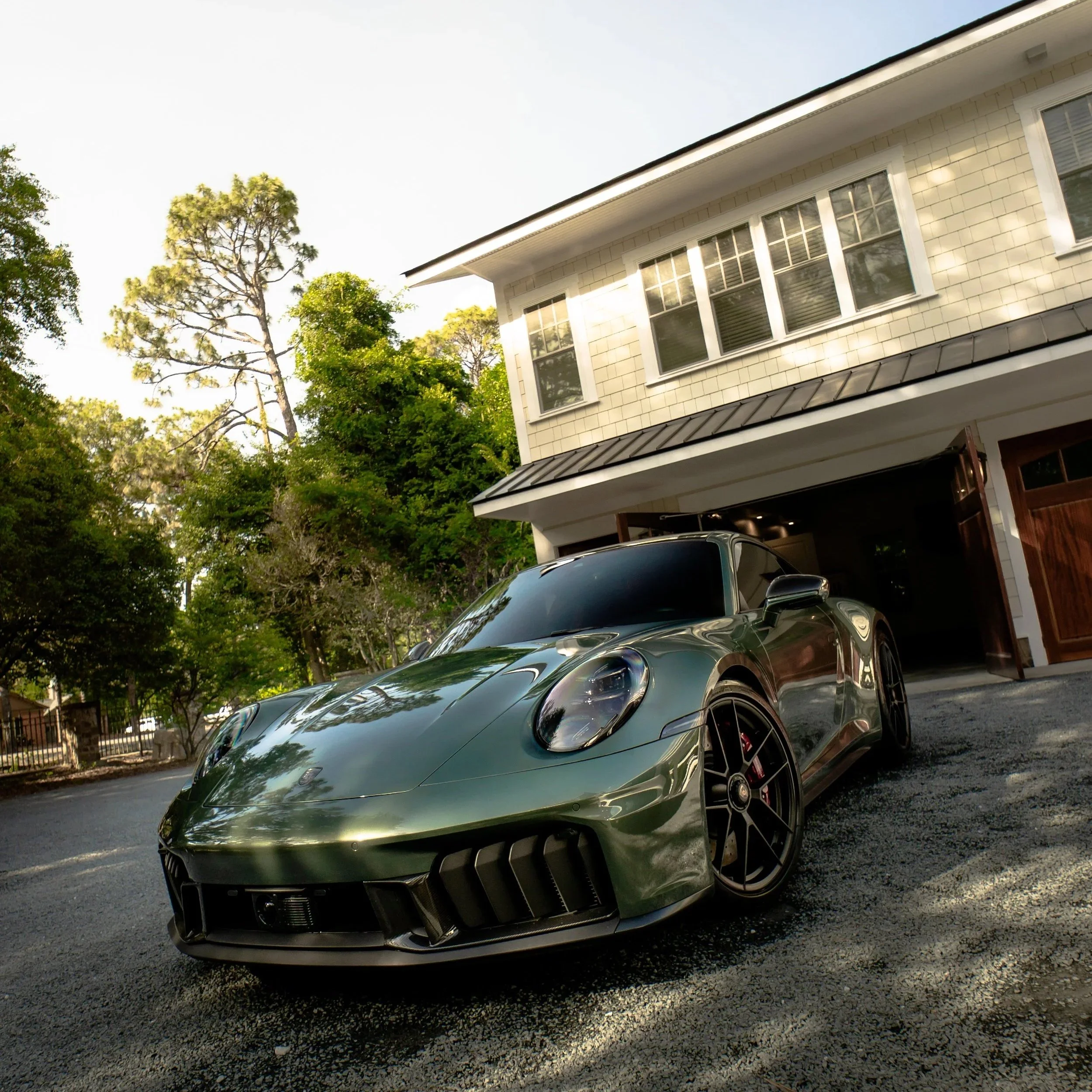 Emerald green luxury sports car parked in front of a beige house with large windows, surrounded by trees and greenery.