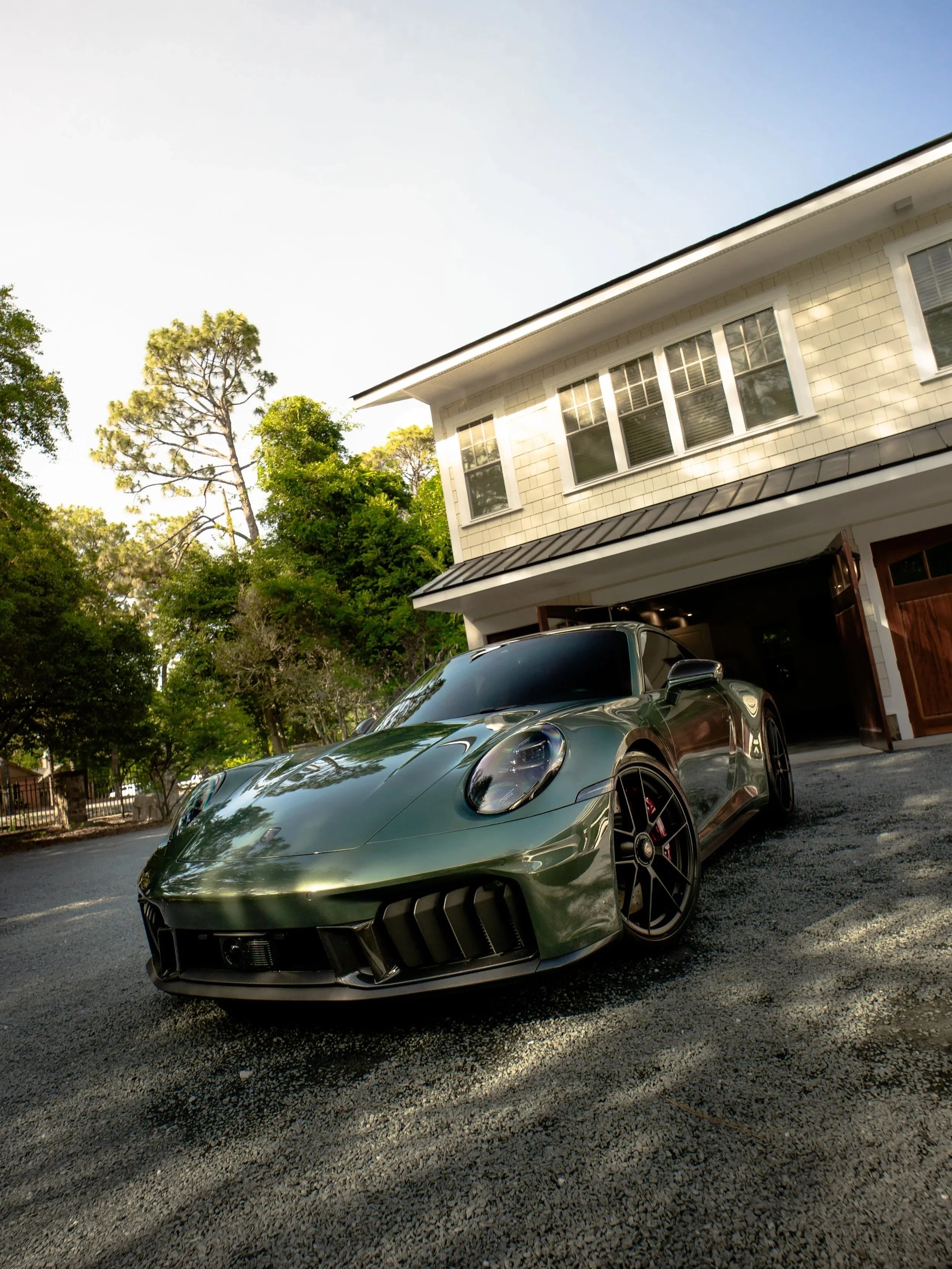 A dark gray sports car parked in a driveway in front of a beige house with white trim and multiple windows, surrounded by green trees. The photo is taken in daylight with a clear sky.