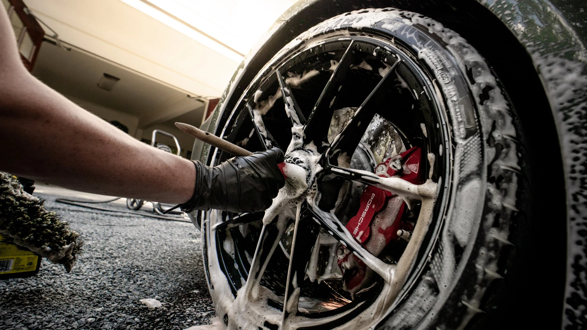 A person washing a black sports car with foam and soap suds, focusing on the wheel and brake caliper.