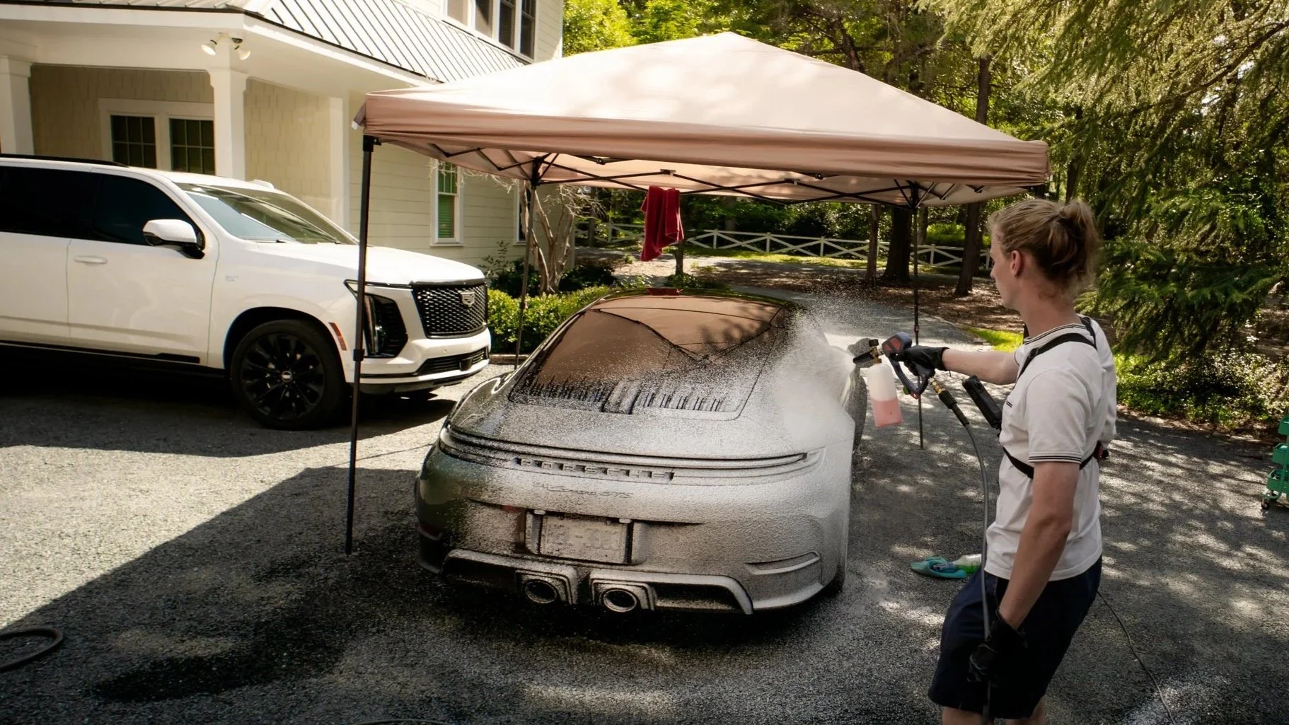 A person is washing a silver Porsche sports car with foam using a pressure washer under a canopy, parked on a driveway with a white SUV and a house in the background.