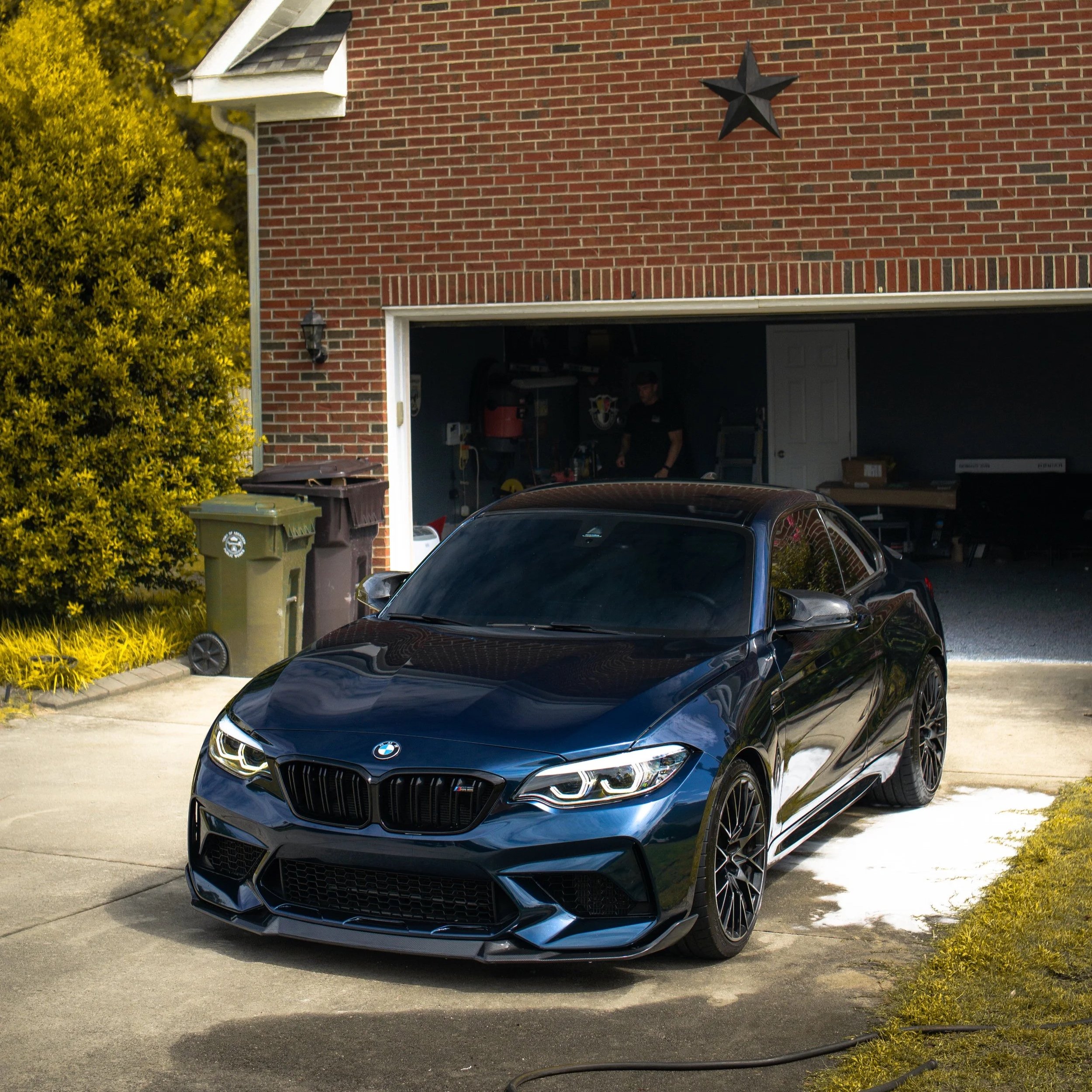 A black BMW sports car parked in front of a brick garage with a star decoration on the wall. The garage appears to be open, with some items visible inside, and there are trees and bushes nearby.
