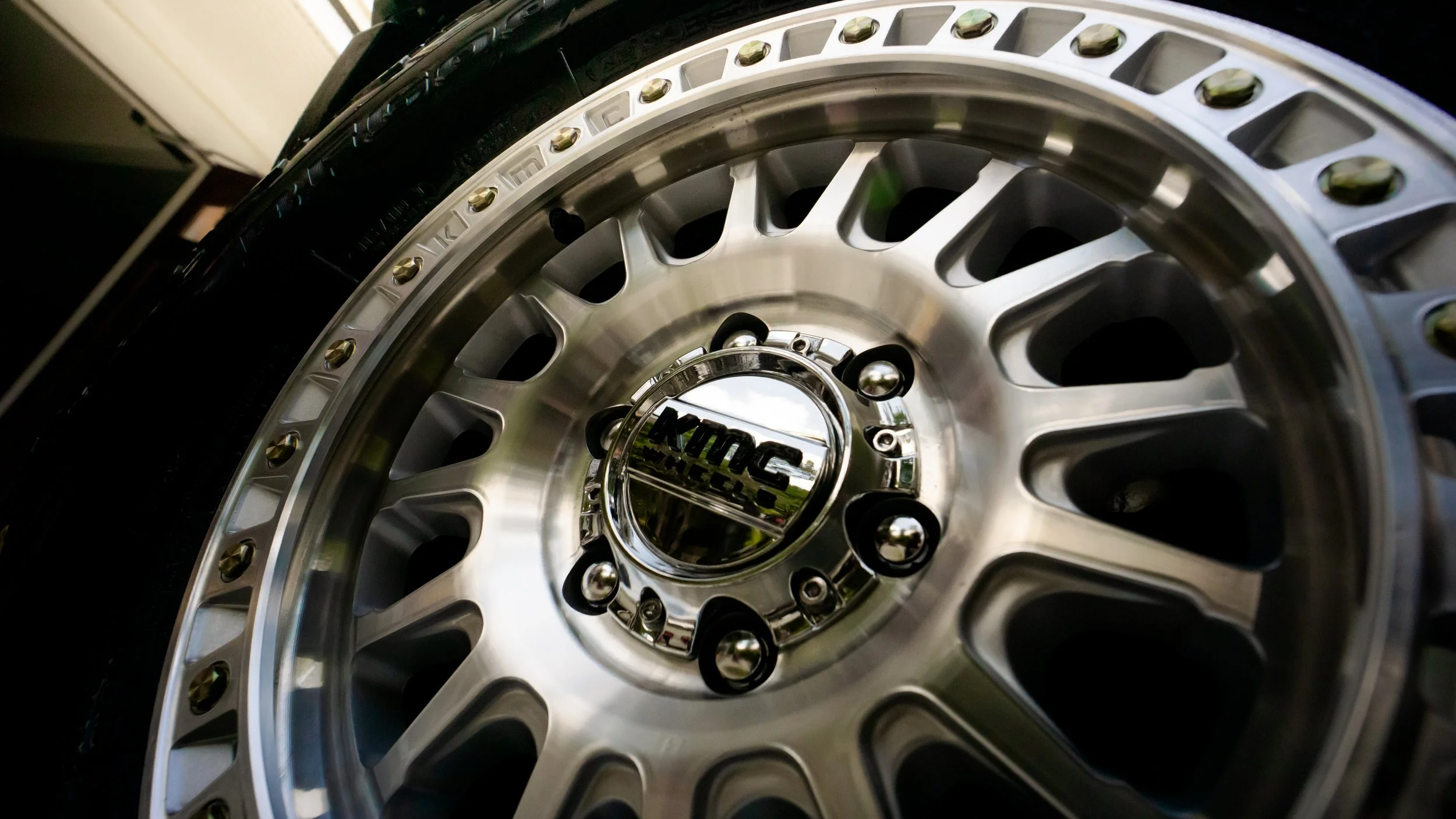 Close-up of a silver aftermarket wheel rim with multiple lug nuts, brand engraved in the center cap, and a black tire mounted on it.