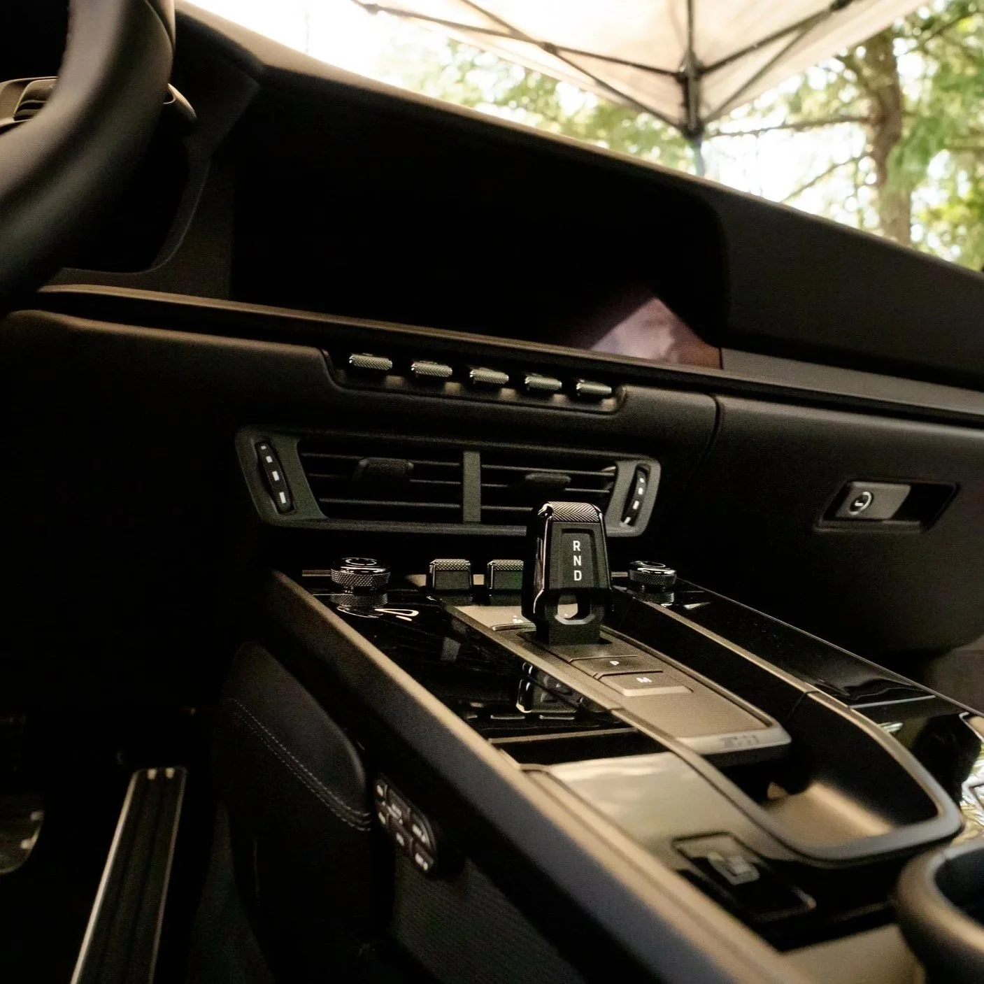 Interior of a modern car showing the gear shift with 'R,' 'N,' and 'D' labels, climate control vents, and a central control panel. Blurred trees are visible outside the windshield.