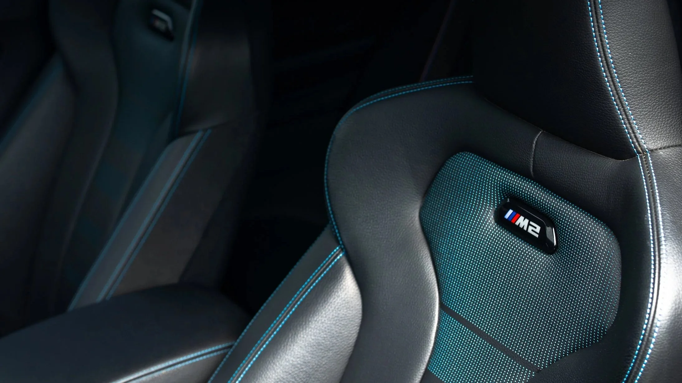 Close-up of a car seat with black leather and blue stitching, featuring an M2 badge on the headrest.