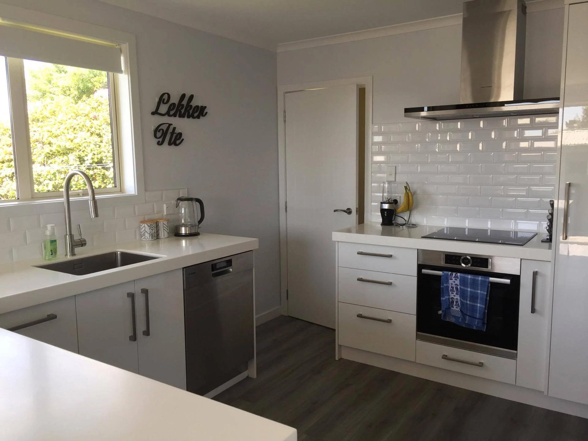 Modern white kitchen with a window looking outside, labeled 'Lakker Ite' on the wall, stainless steel appliances, a black blender, bananas, and a dish towel hanging on the oven handle.