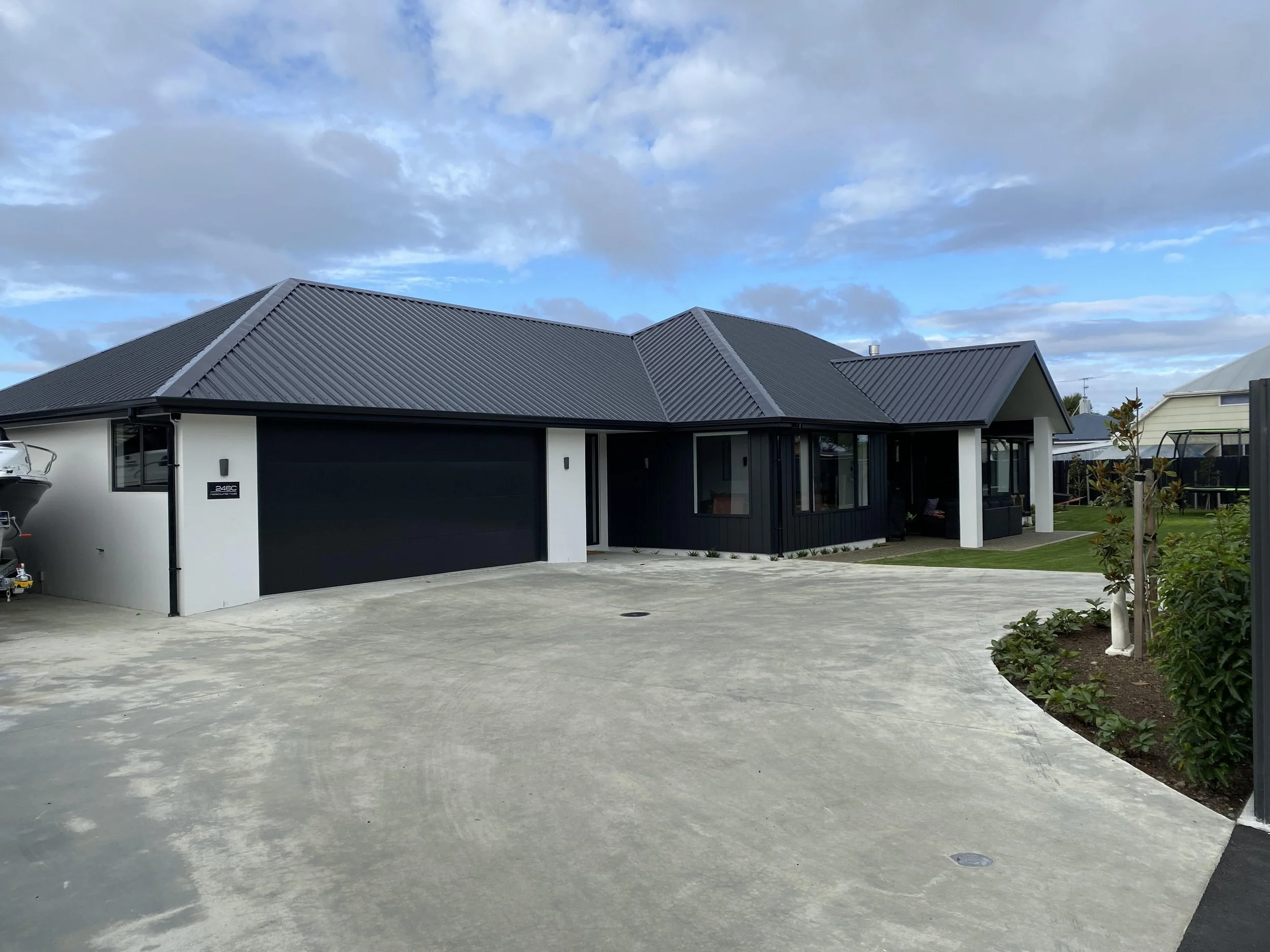 A modern house with a dark metal roof, white walls, and large windows. There is a driveway in front of the house with a car on the left side. The sky is partly cloudy with blue patches. There are some plants and a small garden bed on the right side.