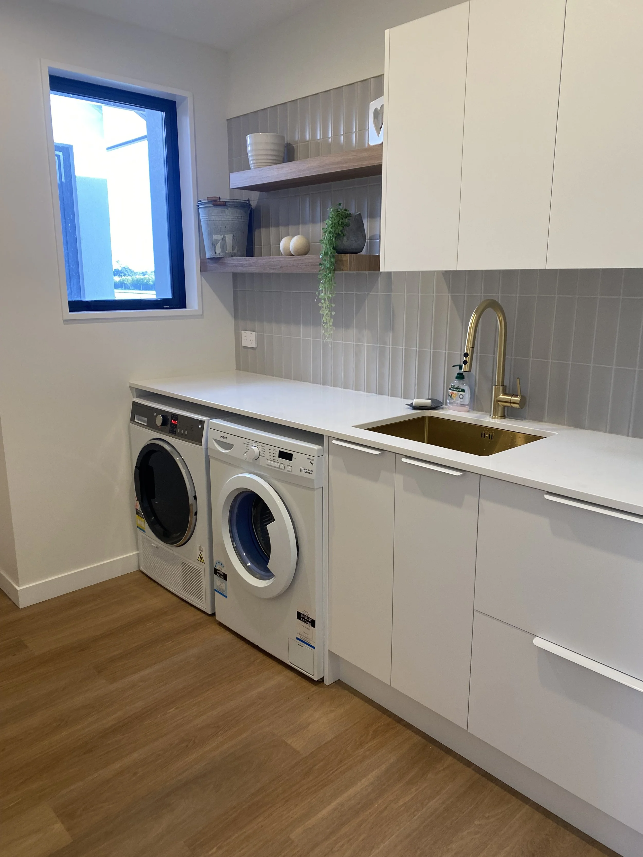 Laundry room with a washing machine and dryer under white cabinets, a sink with a gold faucet, a window, wooden shelves with decoration, and a beige tiled wall.