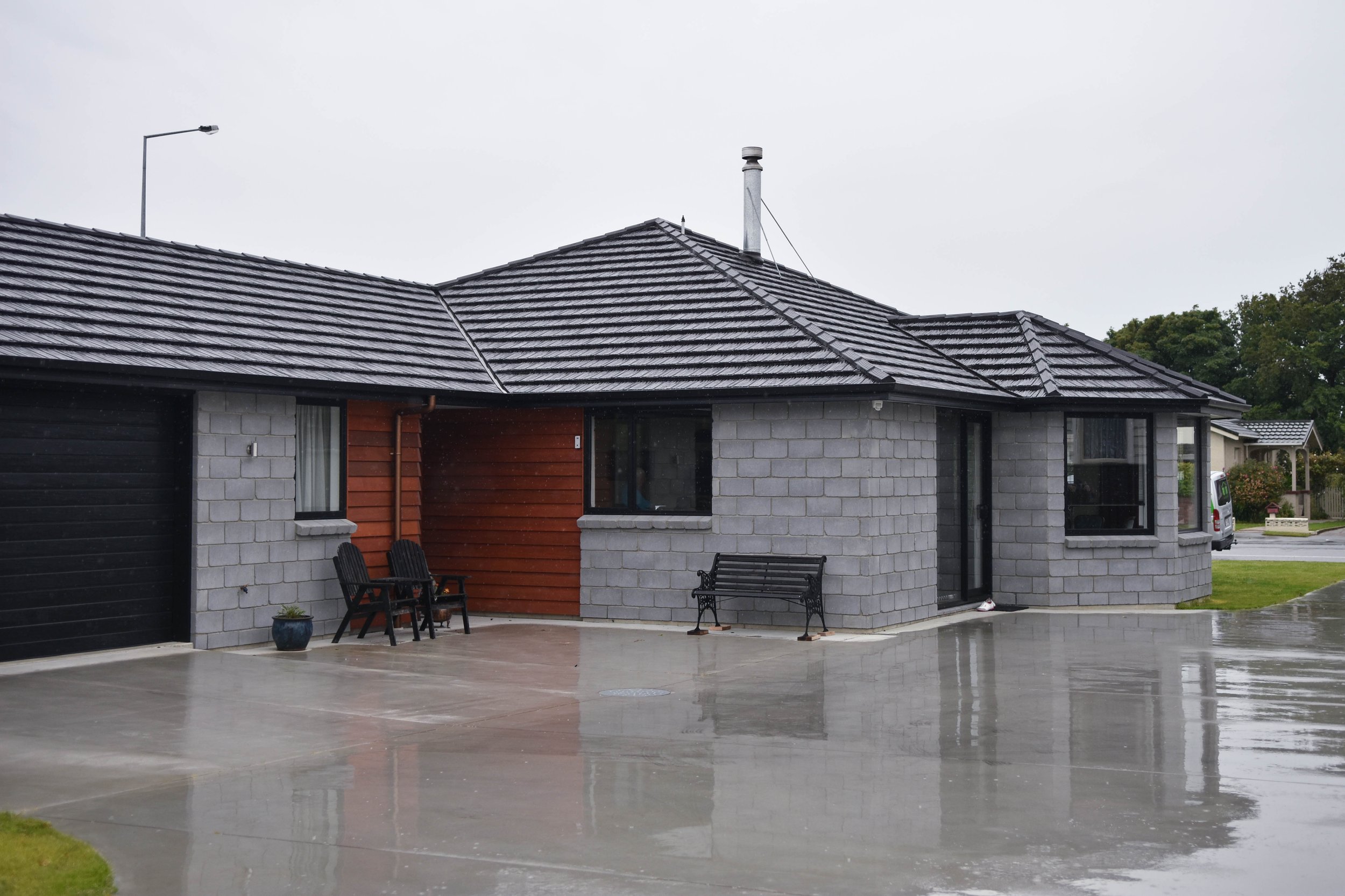A house with a tiled roof, a brick and wood exterior, large windows, and outdoor seating, with wet pavement indicating recent rain.