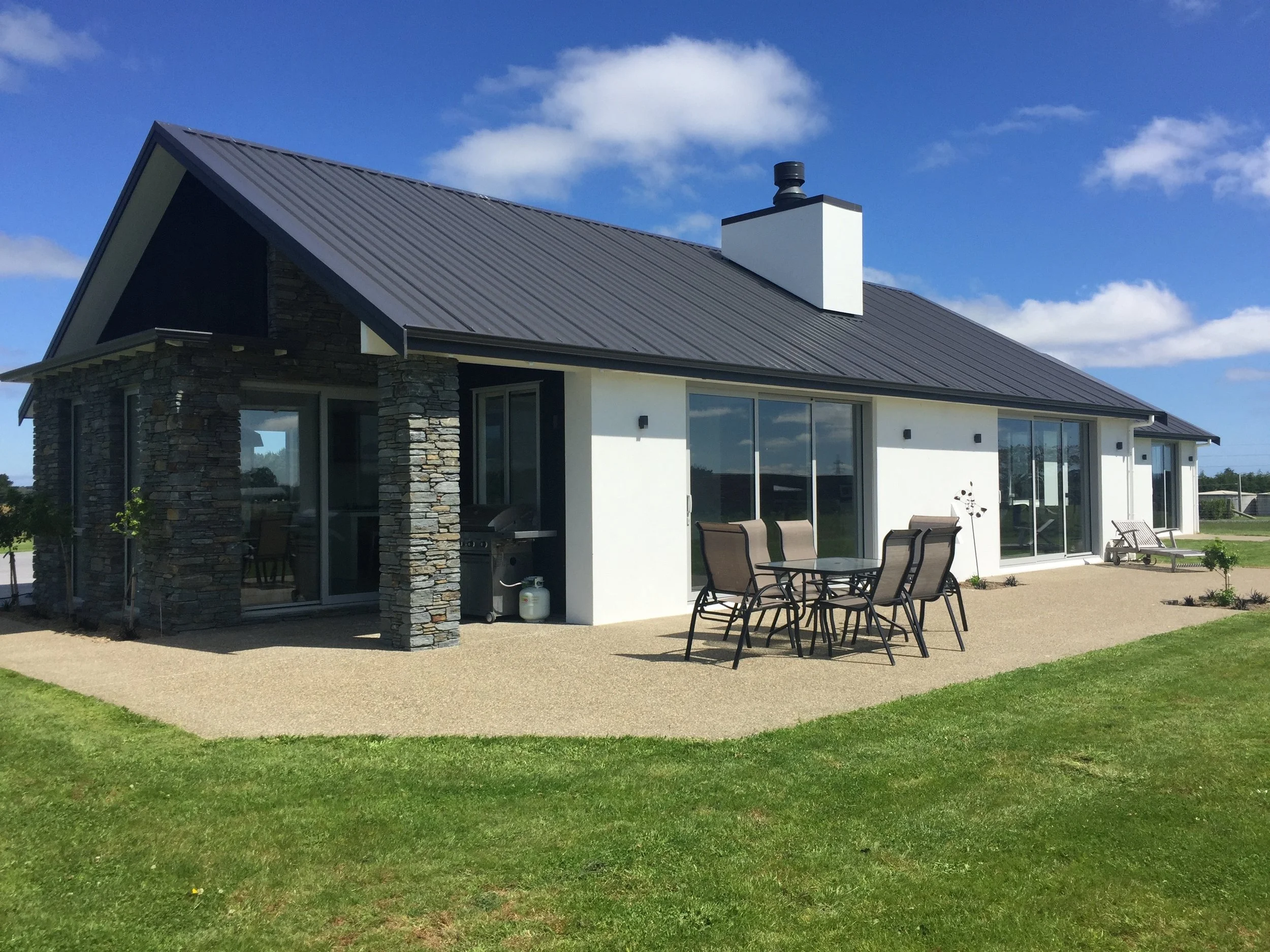A modern house with a sloped metal roof, stone accents, large windows and sliding doors, outdoor patio furniture, and a surrounding well-maintained lawn under a blue sky with some clouds.