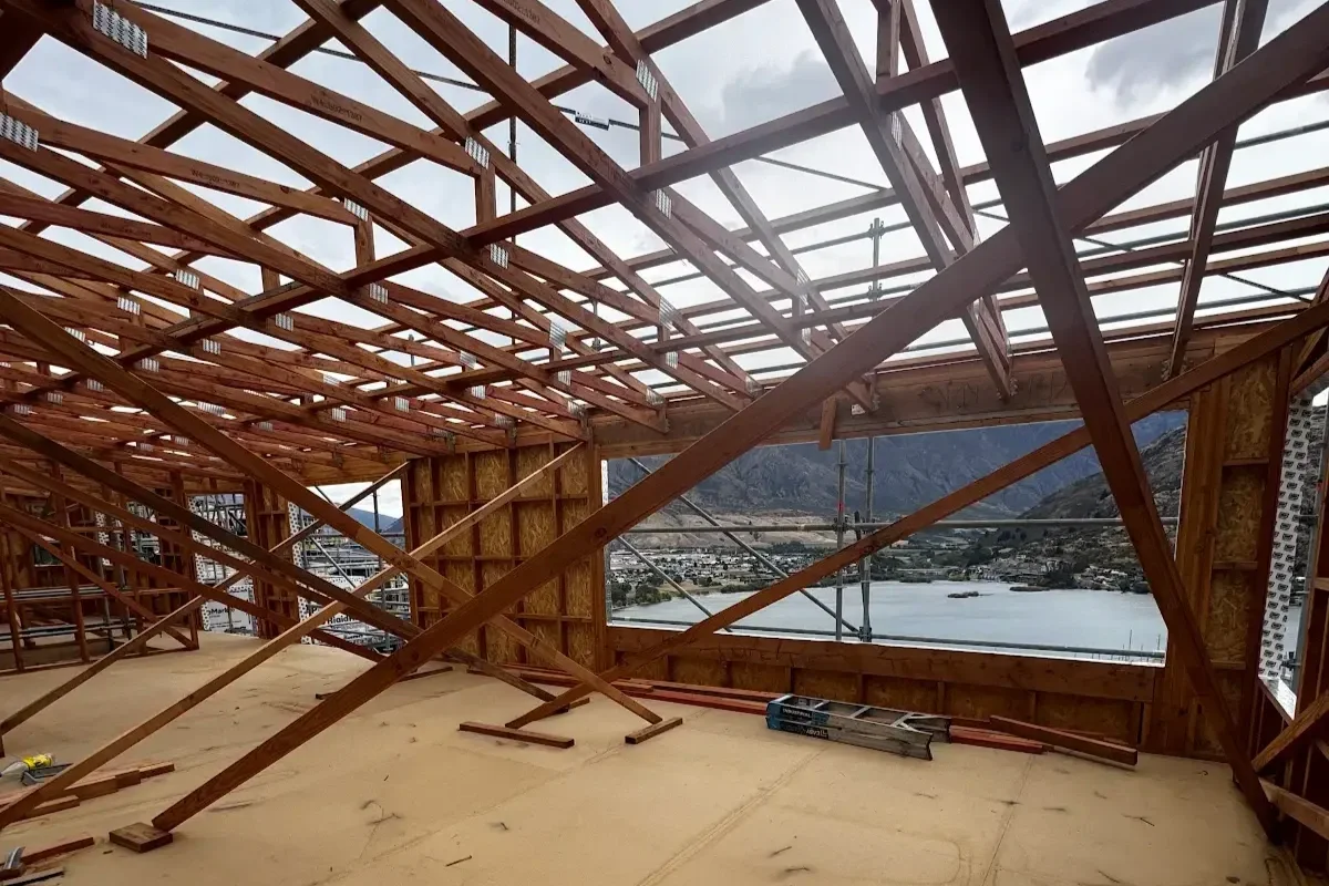 View of a house under construction with exposed wooden framing, scaffolding outside, and a large window revealing a mountain lake landscape.