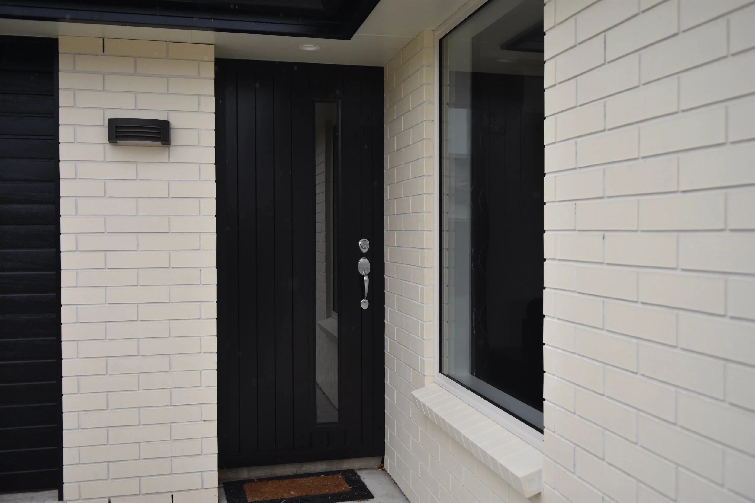 Front entrance with a black door, large windows, cream-colored brick walls, and a black wall-mounted mailbox.