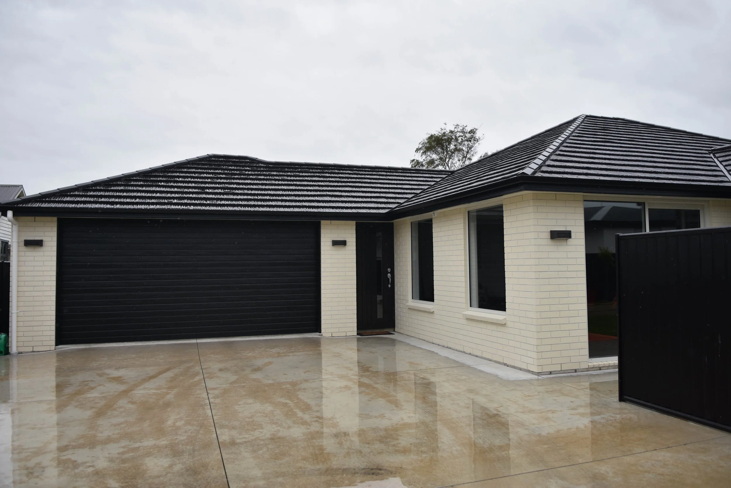 A modern house with a black tiled roof, white brick walls, large windows, and a black garage door, situated on a wet concrete driveway on a cloudy day.