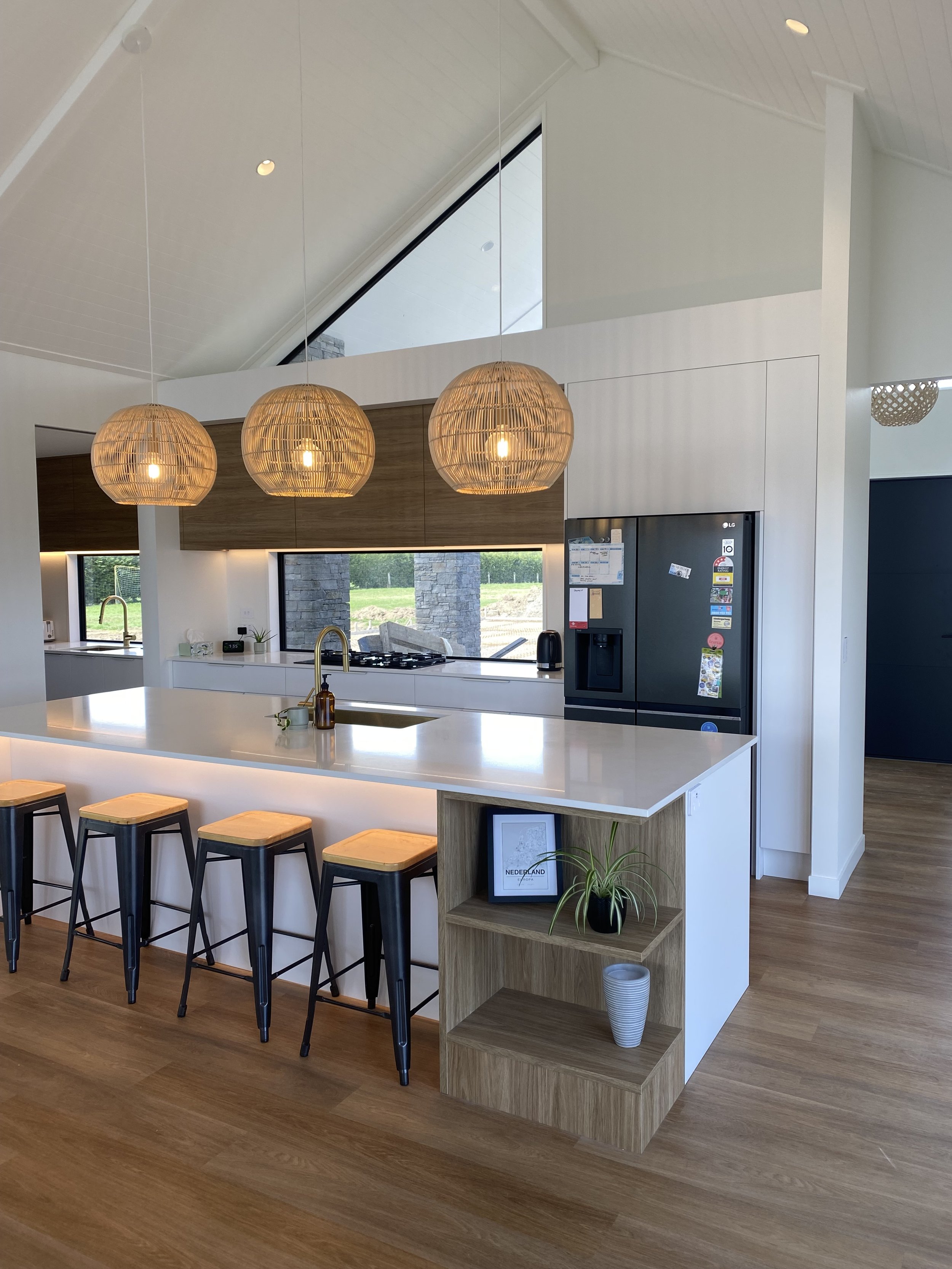 Modern kitchen with wooden bar stools, pendant lights, a white countertop, a black refrigerator, and large windows showing a green outdoor area.