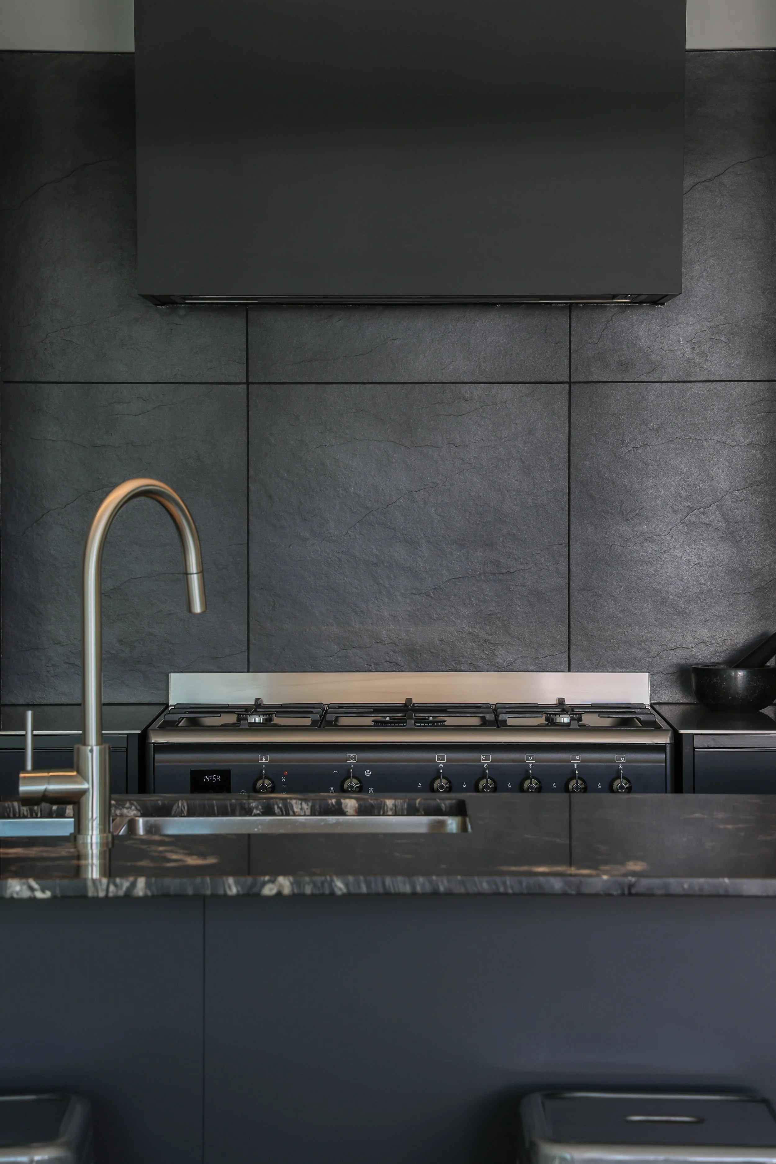 A modern kitchen with a dark gray backsplash, a stainless steel sink with a curved faucet, a stovetop with control knobs, and a black cabinet above the stove.