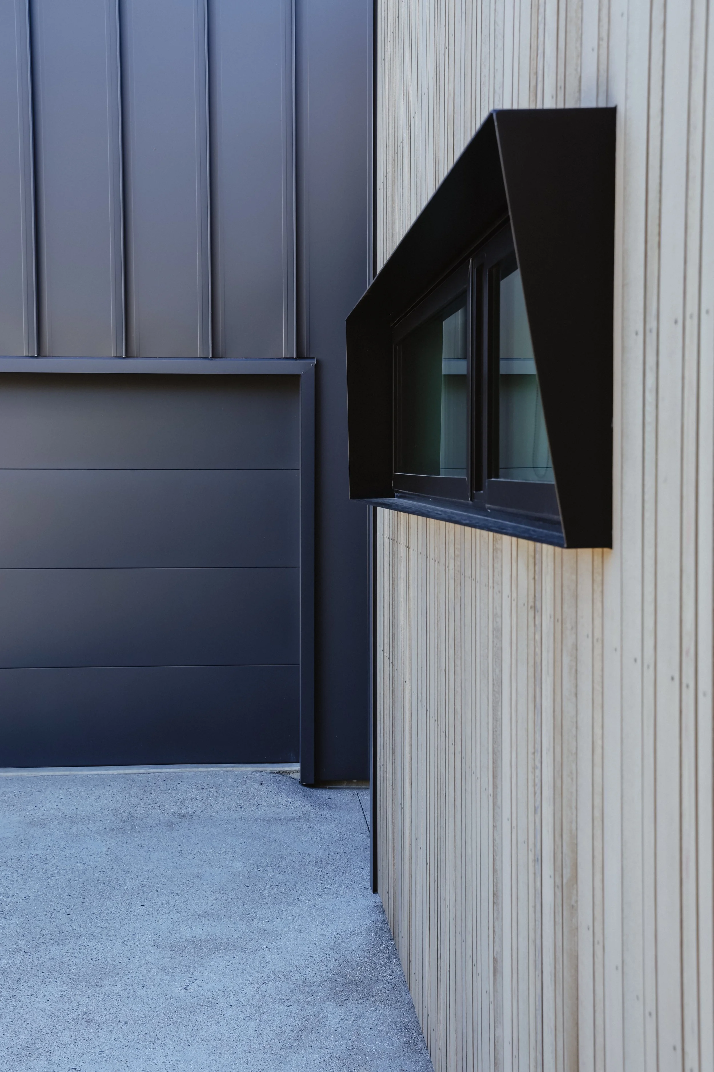 Exterior view of a modern building with dark gray metal siding and light wood paneling. A black-framed window is partially visible on the right side.
