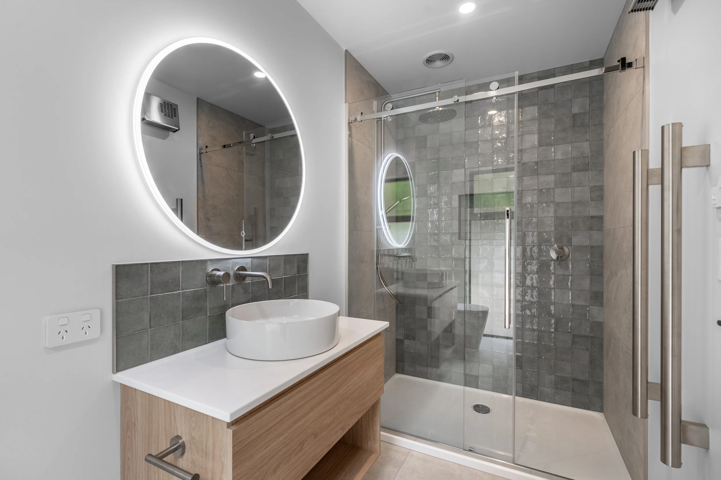 Modern bathroom with a round illuminated mirror, a vessel sink on a wooden vanity, a glass shower enclosure with a rainfall showerhead, gray tiled walls, and a window with a view of greenery.