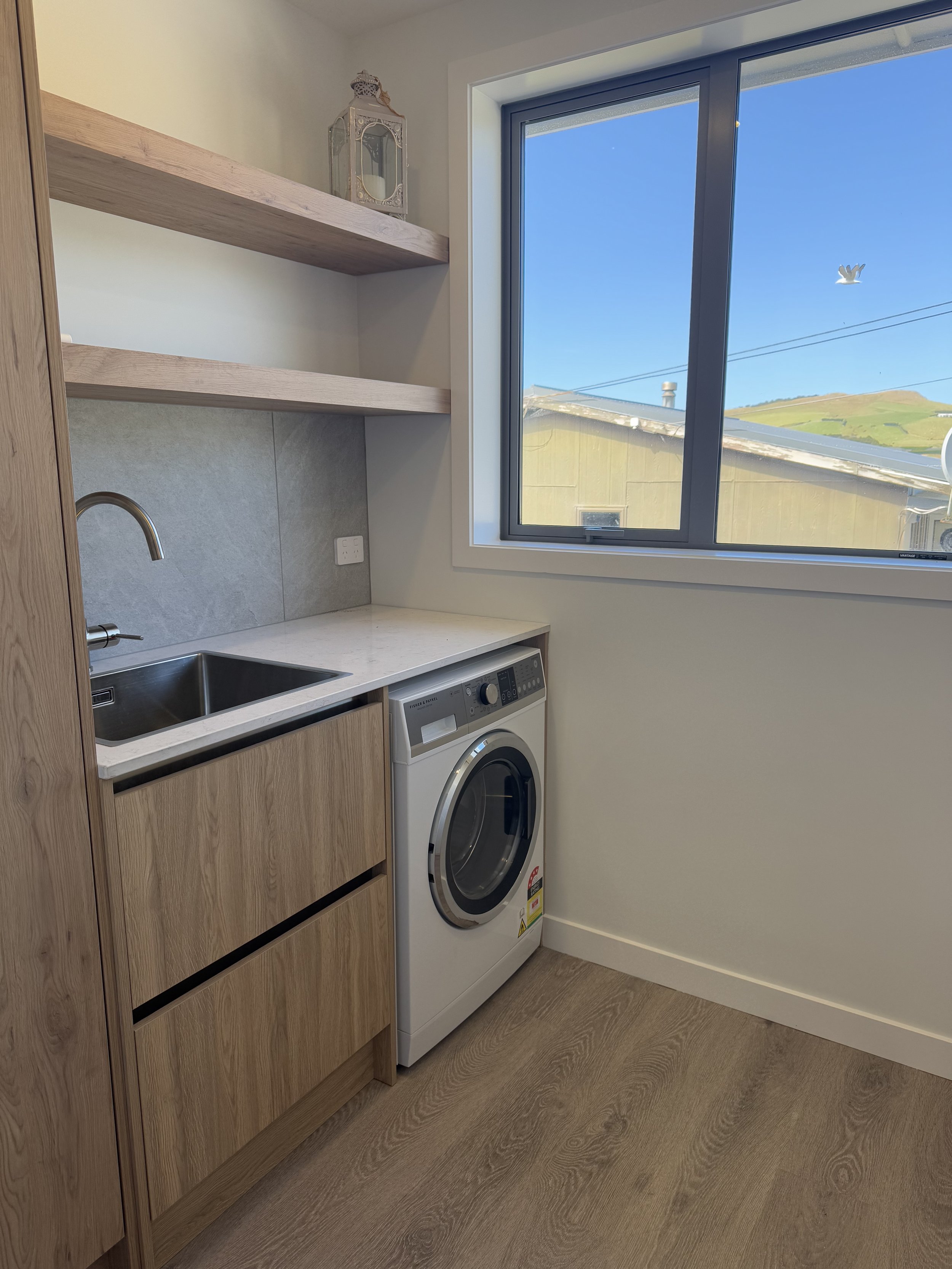 A small laundry room with light-colored wood cabinetry, a stainless steel sink, a washing machine, and a window showing a view of green hills and a blue sky.