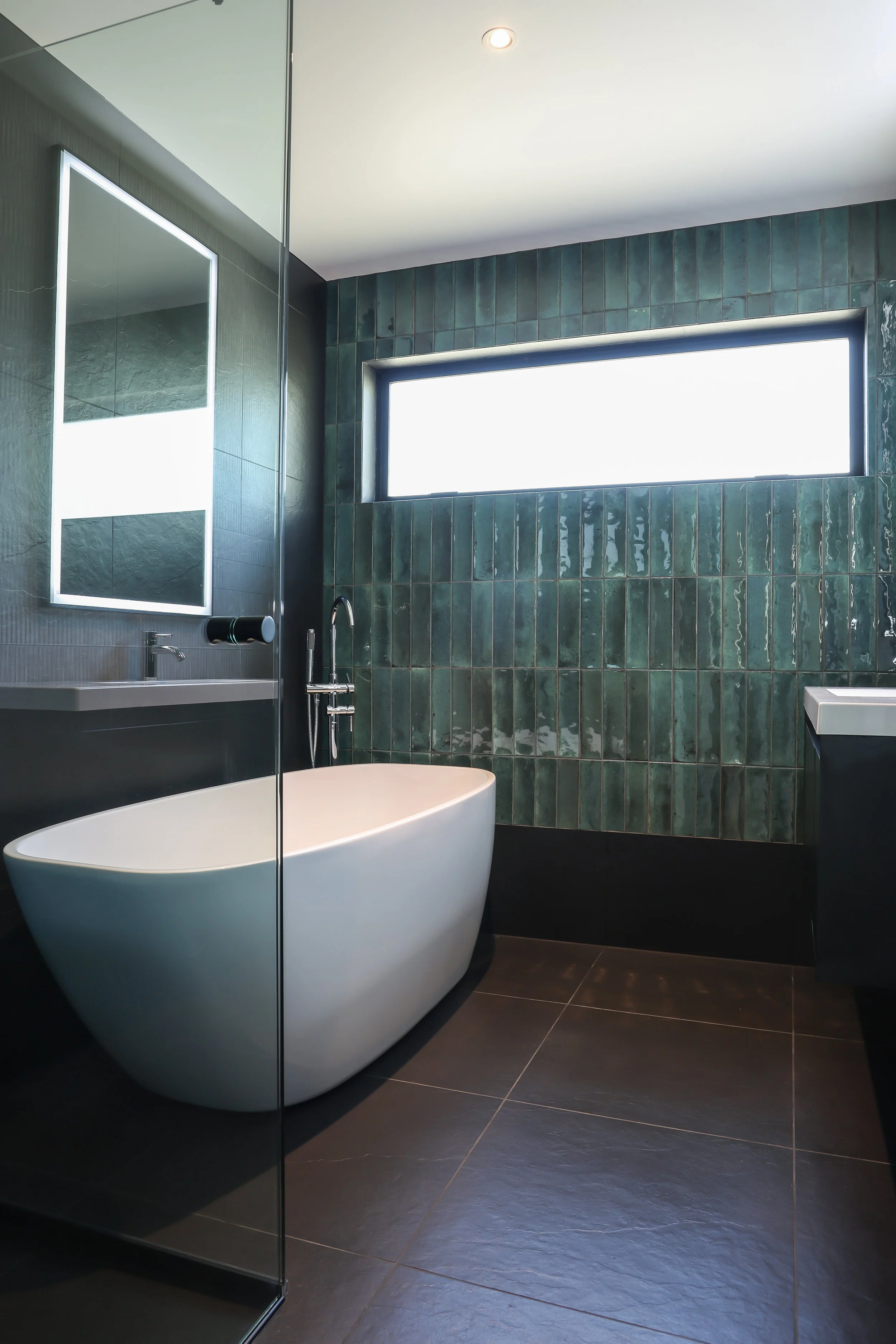 Modern bathroom with a free-standing bathtub, dark tile flooring, a green tiled wall, a rectangular window, and a mirrored cabinet.