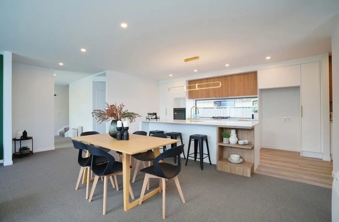 Open-concept living space with a dining area and kitchen, featuring a light wood dining table, black chairs, a modern kitchen island with black barstools, white cabinetry, a window above the sink, and minimalist decor.