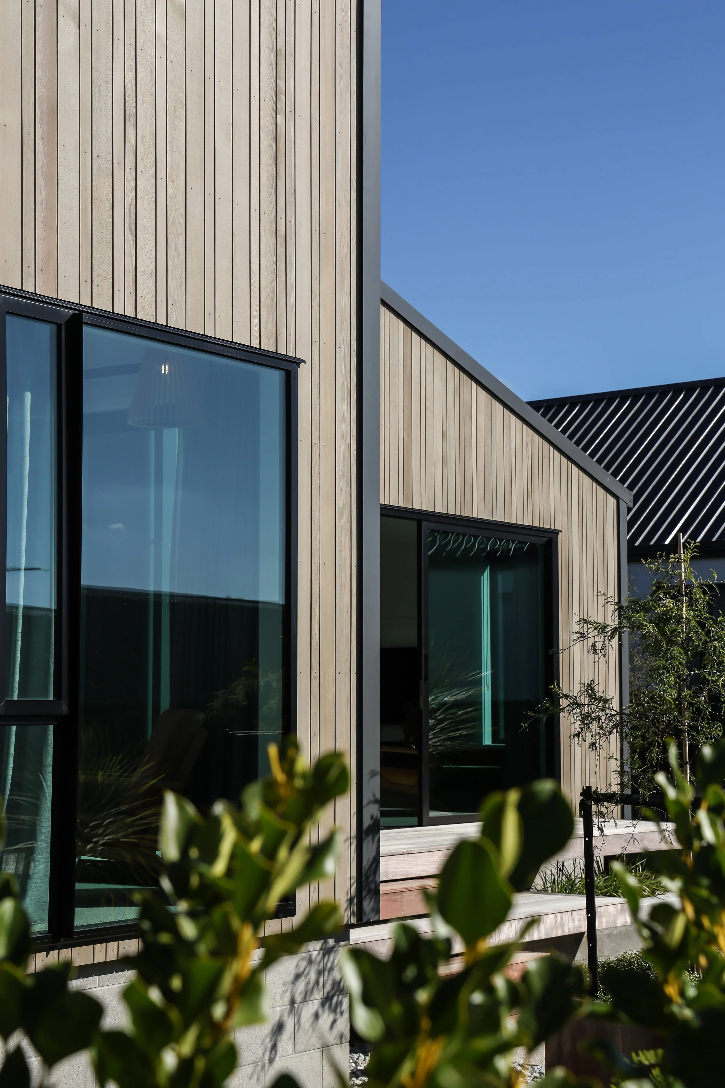 Modern house with wood panel exterior, large glass windows, and a small deck with railing, surrounded by greenery and trees, under a blue sky.