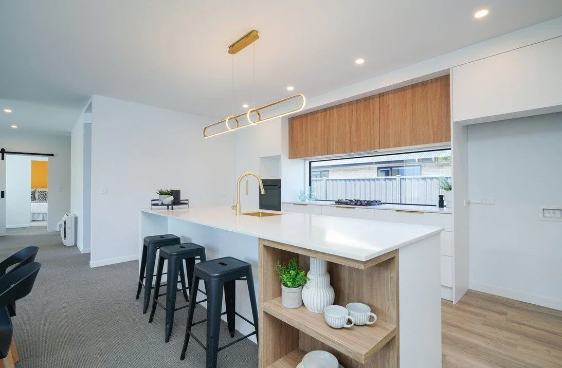Modern kitchen with white countertops, gold fixtures, black bar stools, wooden cabinets, and a large window.