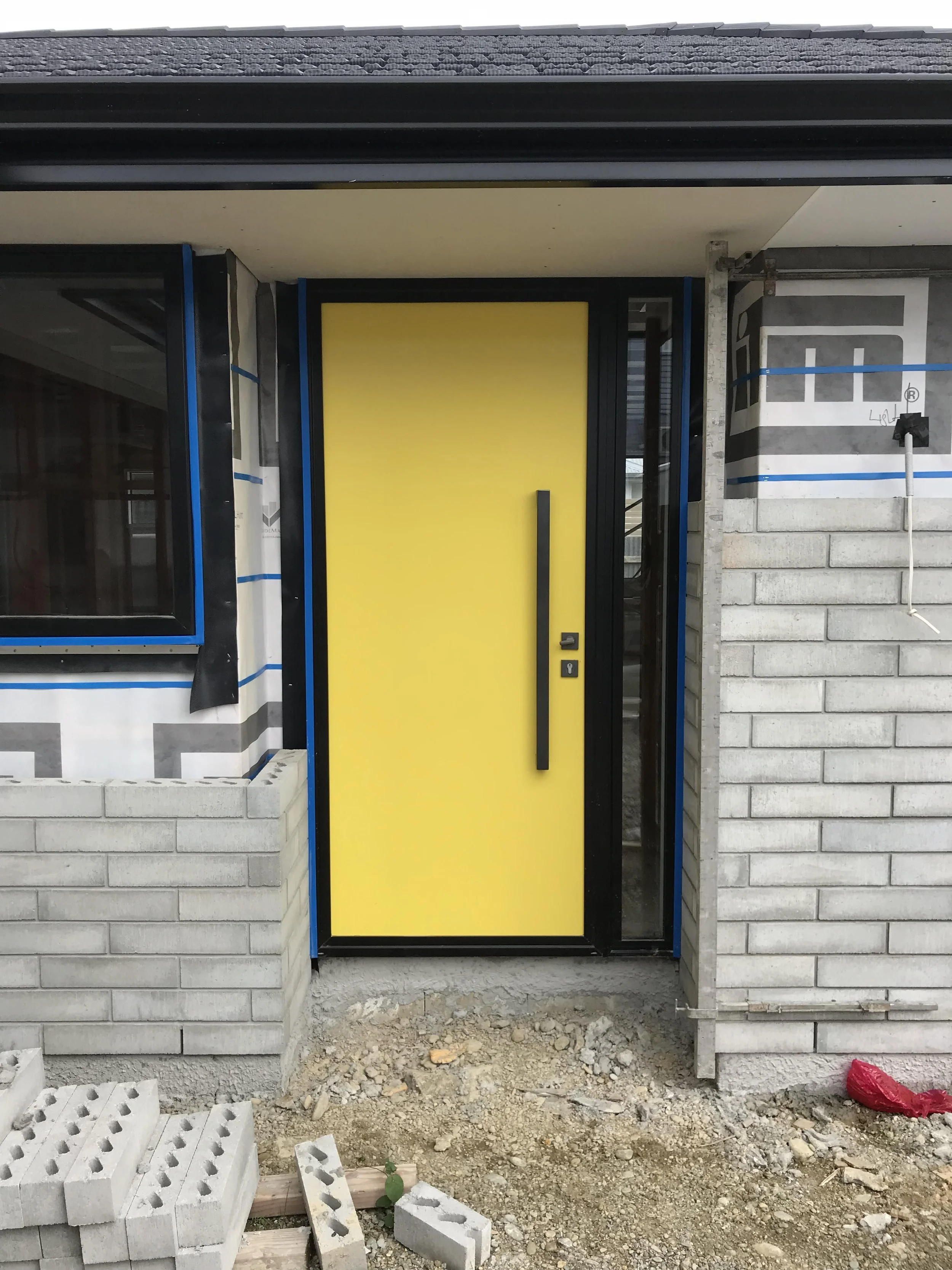 Under construction house entrance with a yellow door, brick walls on each side, construction blocks, and dirt ground in front.