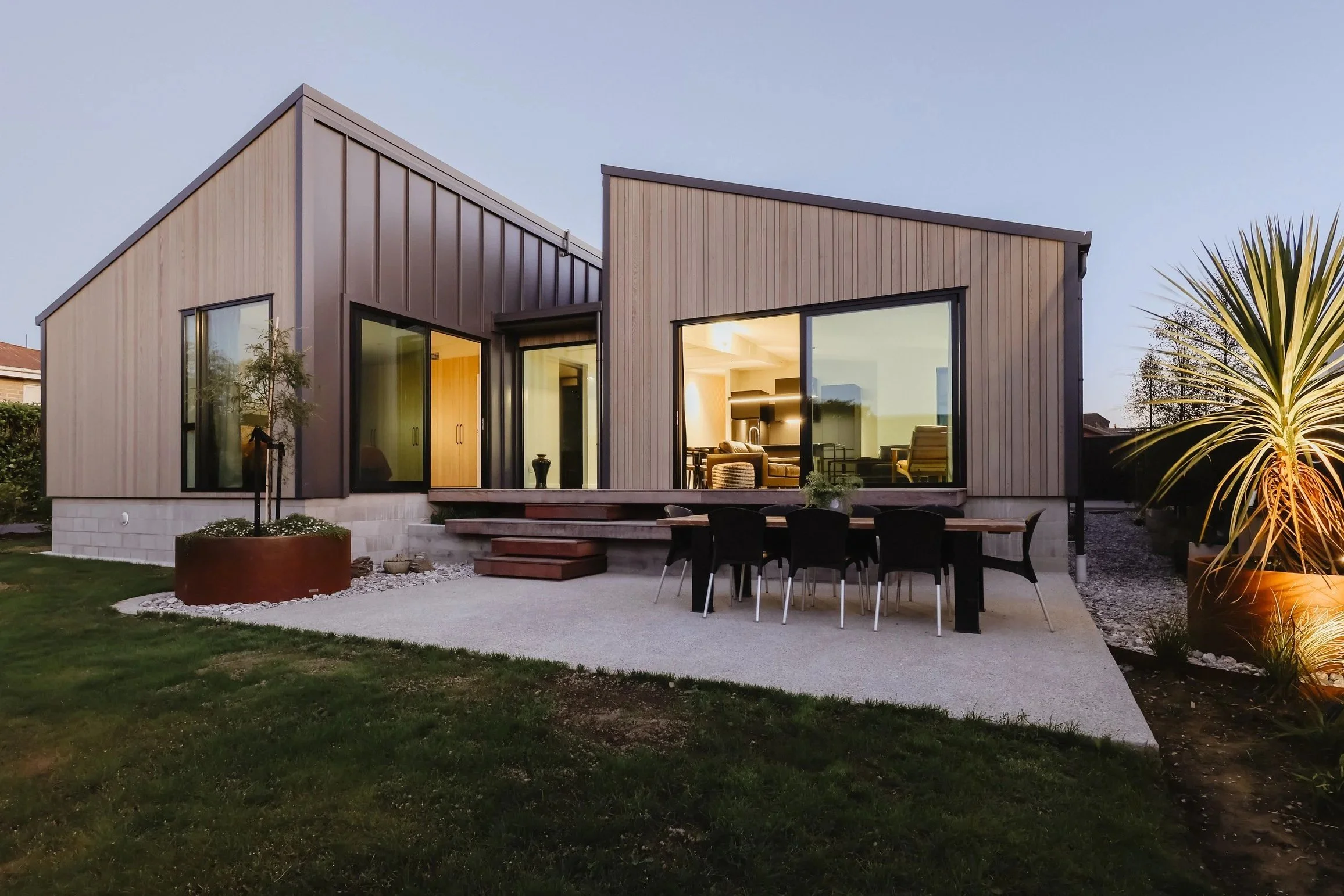Modern house with large glass windows and an outdoor dining area, illuminated from inside during dusk, with a landscaped lawn and potted plants.