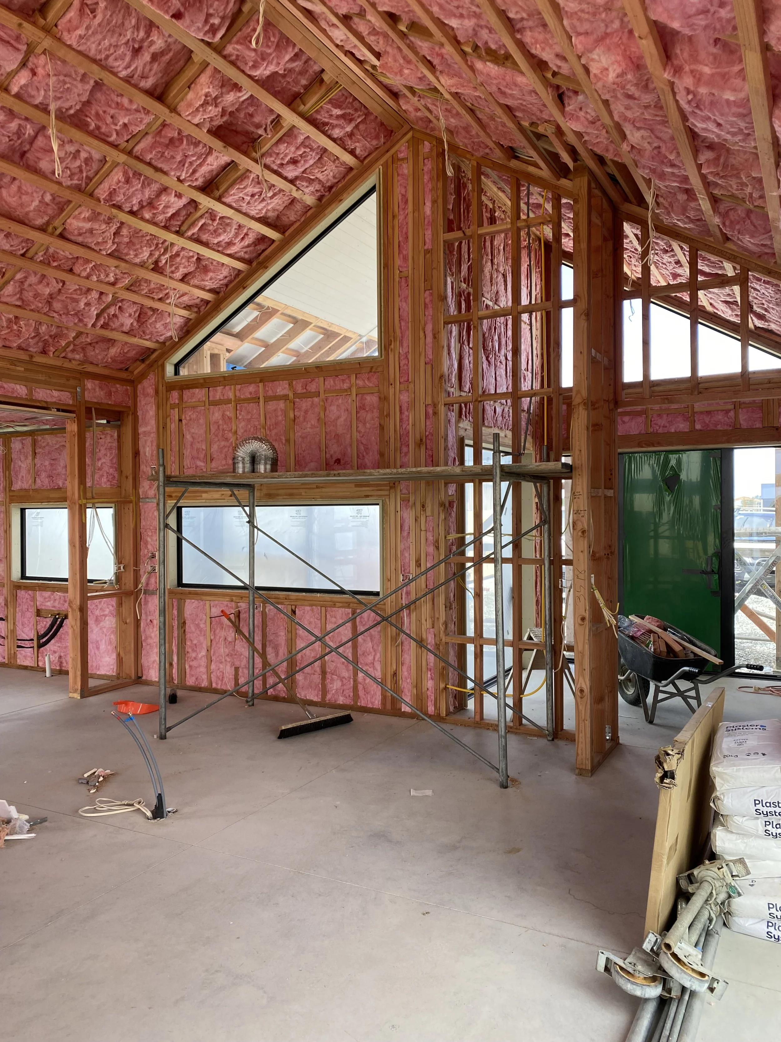 Interior of a house under construction, with exposed pink insulation and wooden framing. There are construction materials, a wheelbarrow, scaffolding, and various pipes and wiring visible.