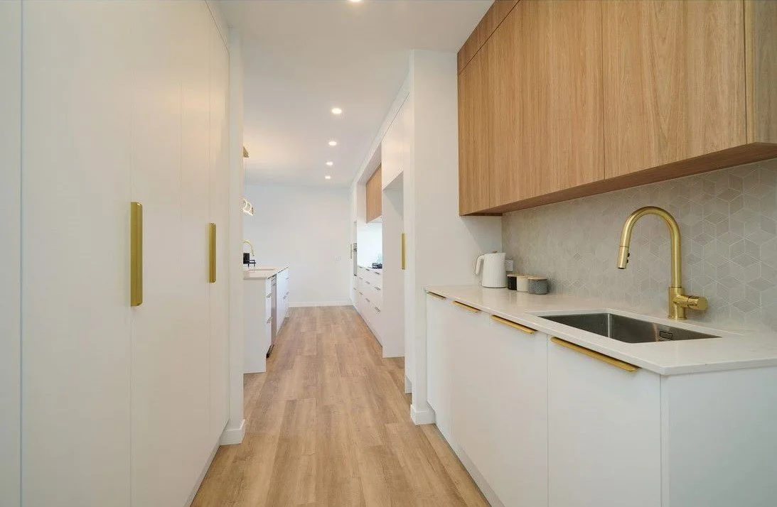 Modern kitchen with white and wood cabinets, gold handles, and a gold faucet. Light wood flooring. Counter with a kettle and containers. Bright, minimalist design.