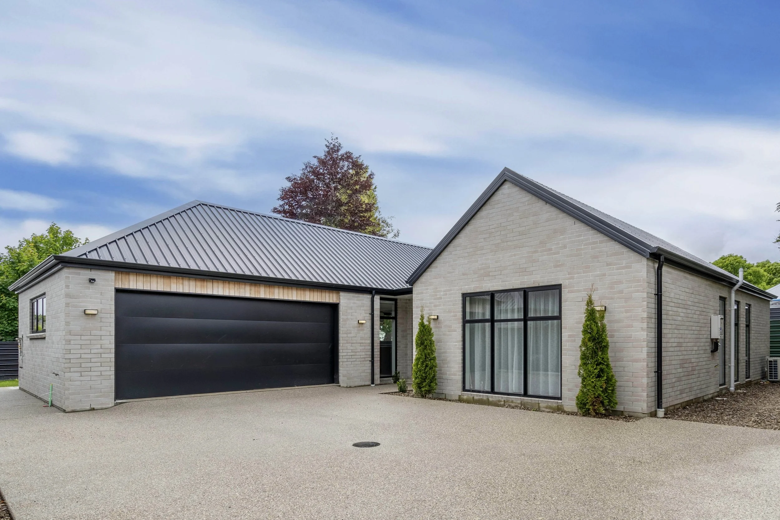 Modern suburban house with light brick exterior, black garage door, large glass windows, small trees in front, and a grey metal roof, under a partly cloudy sky.