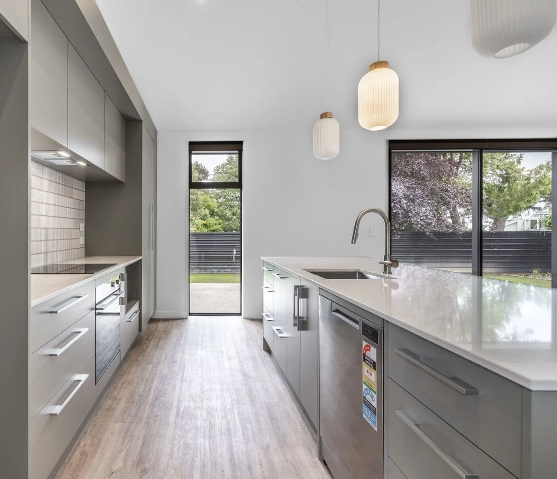 Modern kitchen with white cabinetry, stainless steel appliances, a white island with a sink, pendant lights, and large windows showing greenery outside.