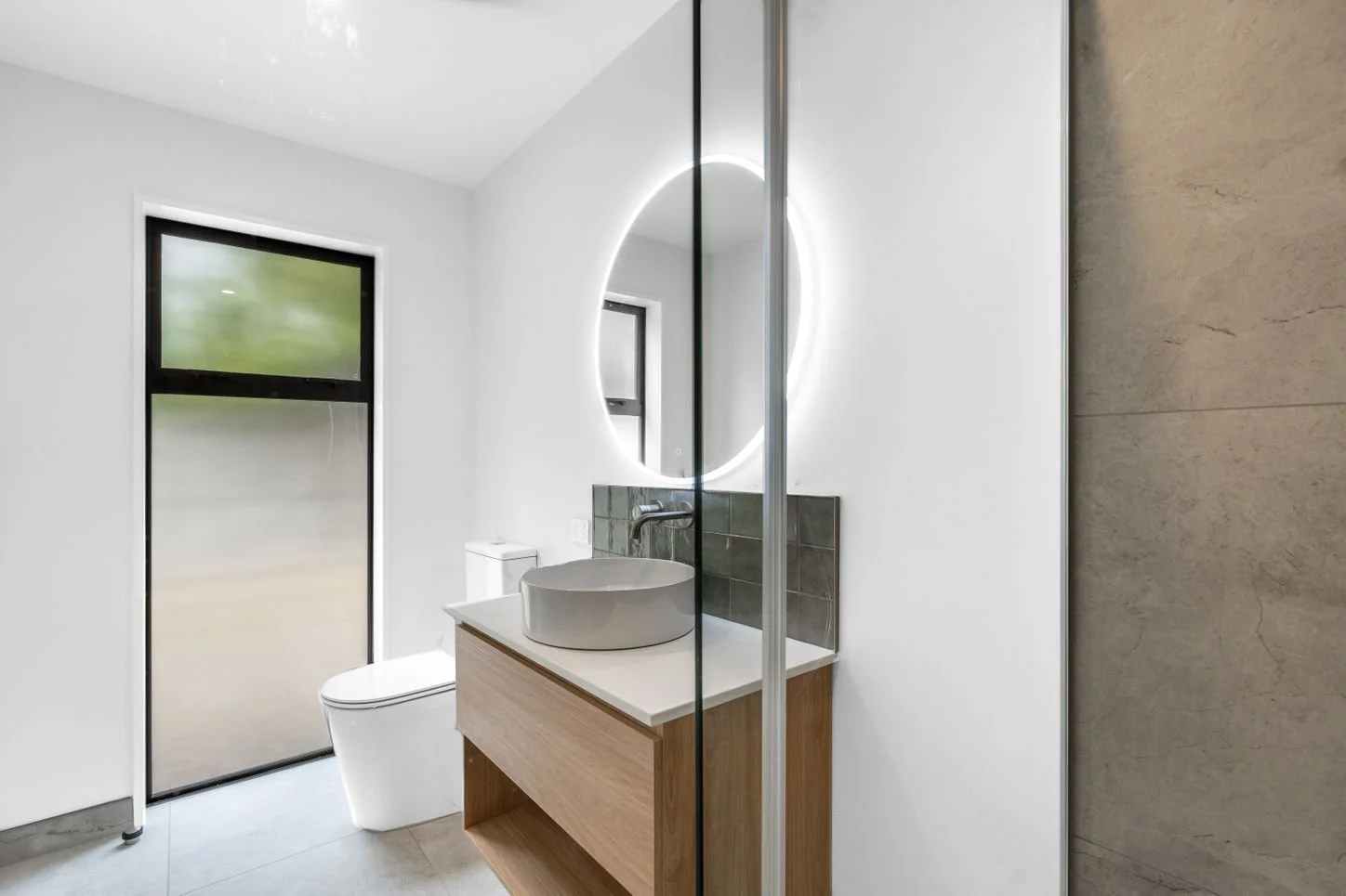 Modern bathroom with round vessel sink, illuminated mirror, frosted glass door, toilet, wooden vanity, and light-colored floor tiles.