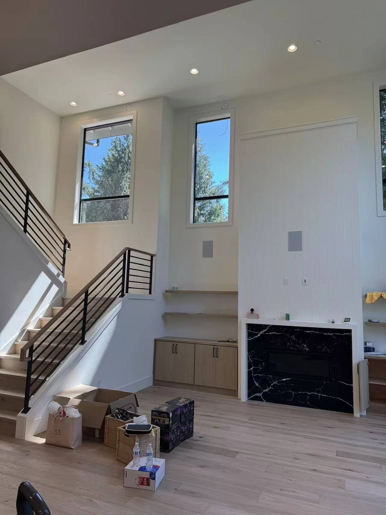 Interior of a modern living space with high ceilings, large windows, staircase with black metal railing, and a partial view of a fireplace with black marble surround. There are boxes, bags, and bottles on the floor, indicating ongoing interior work o
