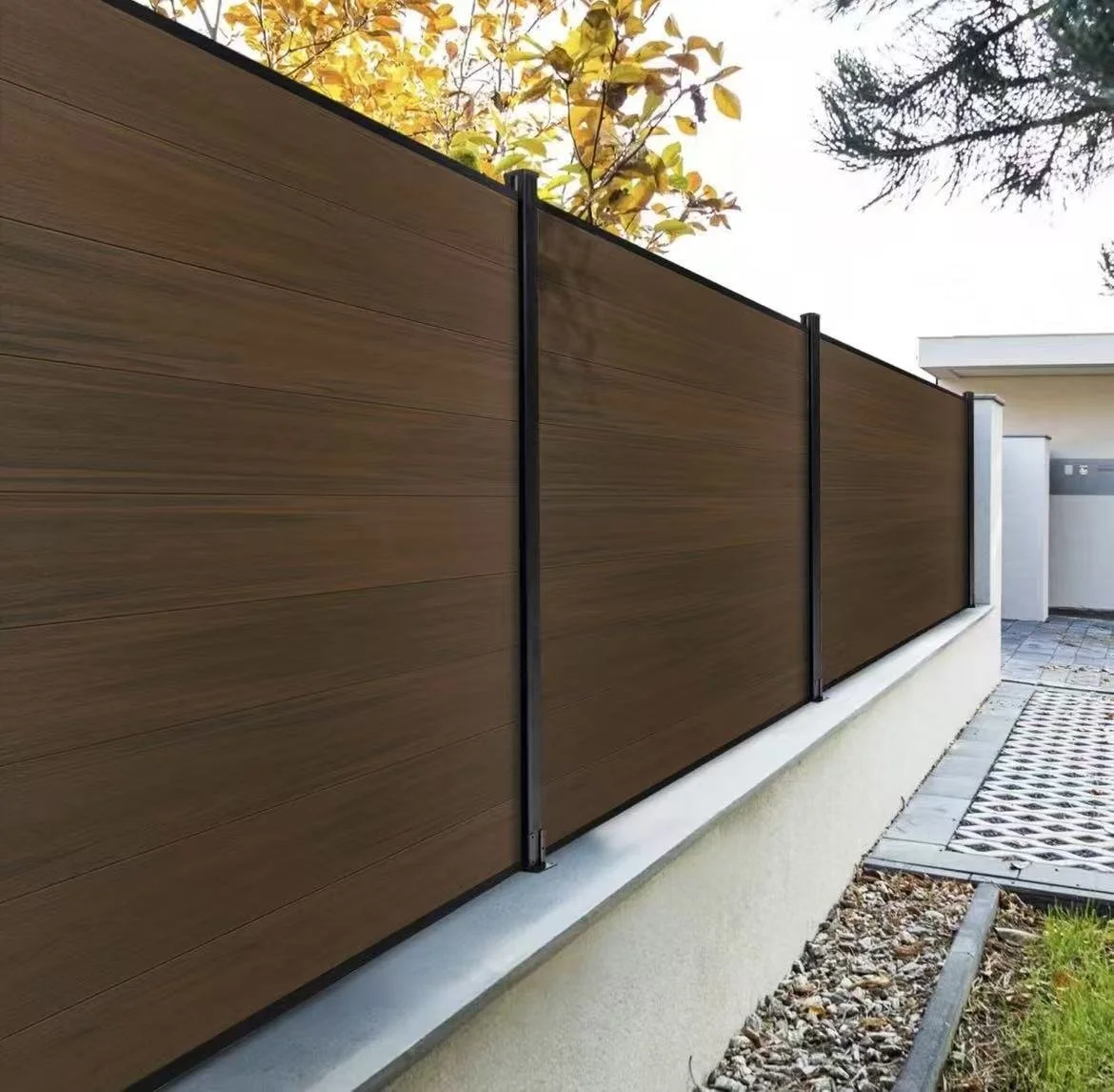 Wooden fence panels with black metal supports along a concrete base, next to a walkway with a metal grate and soil with small rocks, trees with autumn leaves visible in the background.