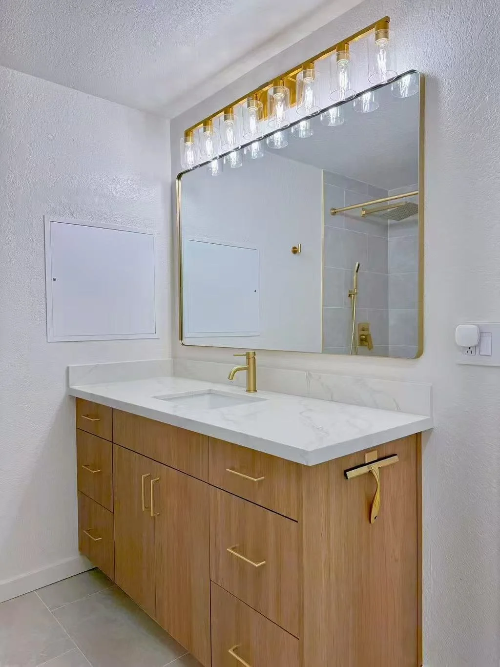 Modern bathroom vanity with a wooden cabinet, white marble countertop, and gold fixtures, reflected in a large mirror with light bulbs on top, showing the shower area with a gold showerhead.