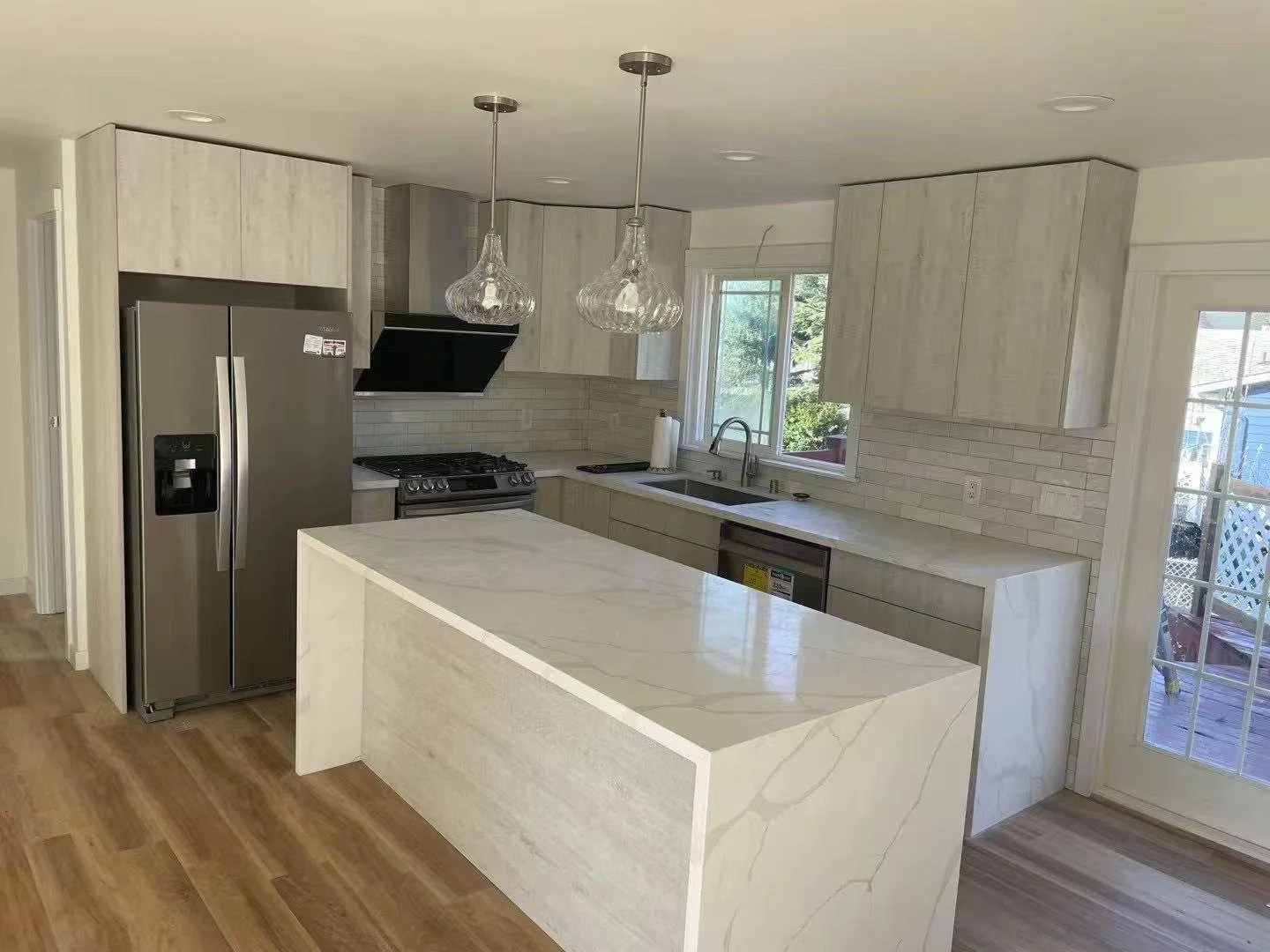 Modern kitchen with white marble island, stainless steel refrigerator, stove, black range hood, white cabinetry, window above sink, and sliding glass door leading to outdoor deck.