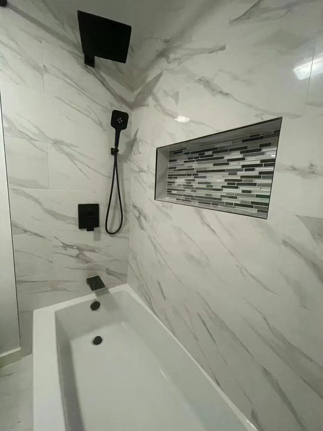 Modern shower with white marble walls featuring gray veining, black fixtures, a rectangular niche with gray and black mosaic tiles, and a white bathtub.