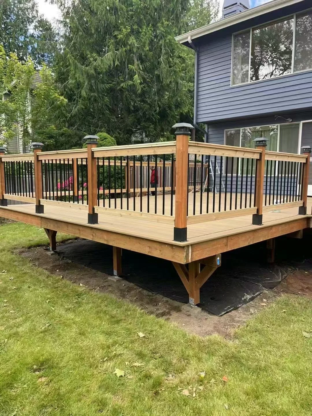 Unfinished wooden deck attached to a blue house with black window trim, surrounded by tall trees and a green lawn.